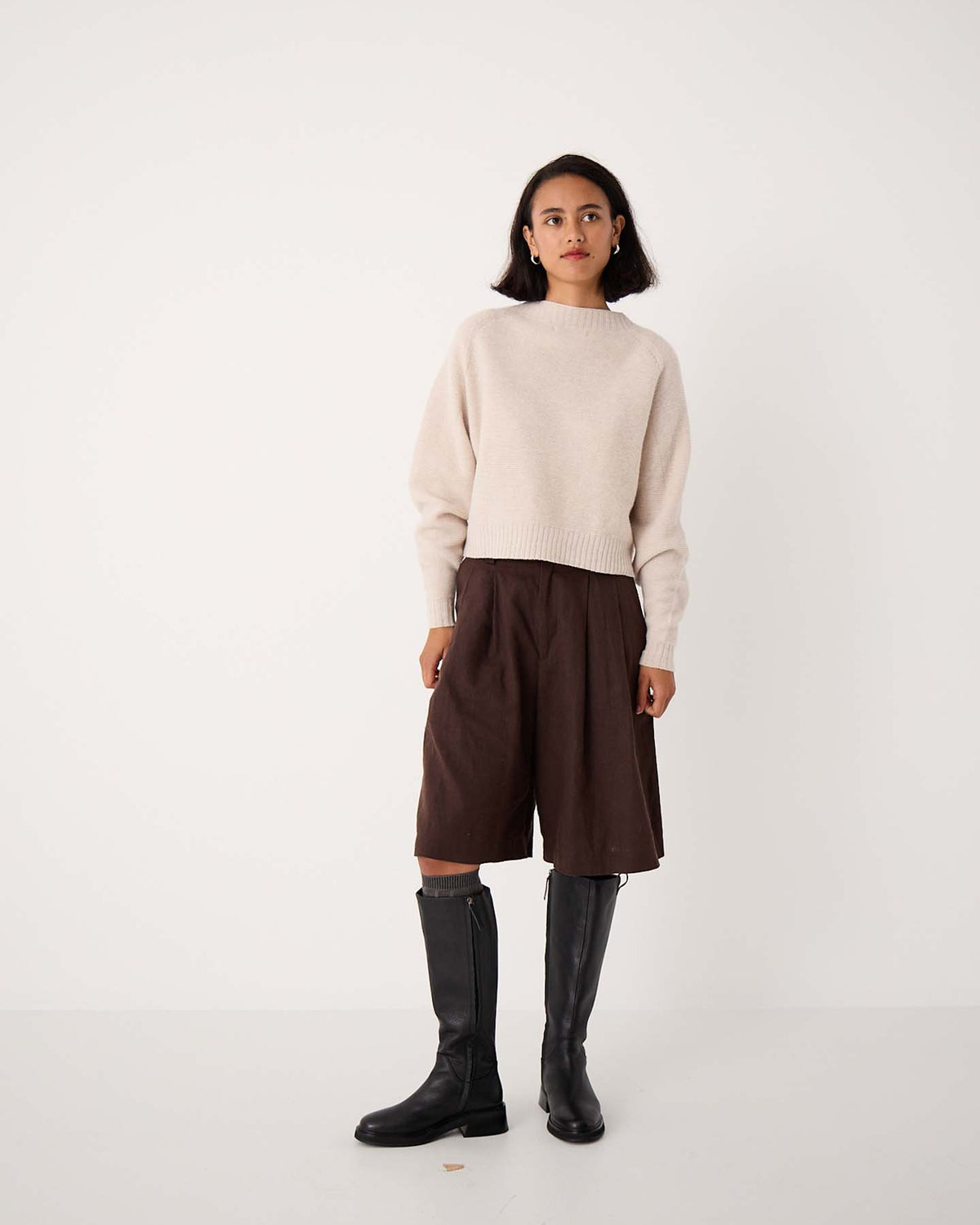 A woman stands against a plain white background wearing a cream sweater, brown knee-length shorts, black knee-high boots, and Tip Toe Knee High Socks in Grey & Cherry Trim.