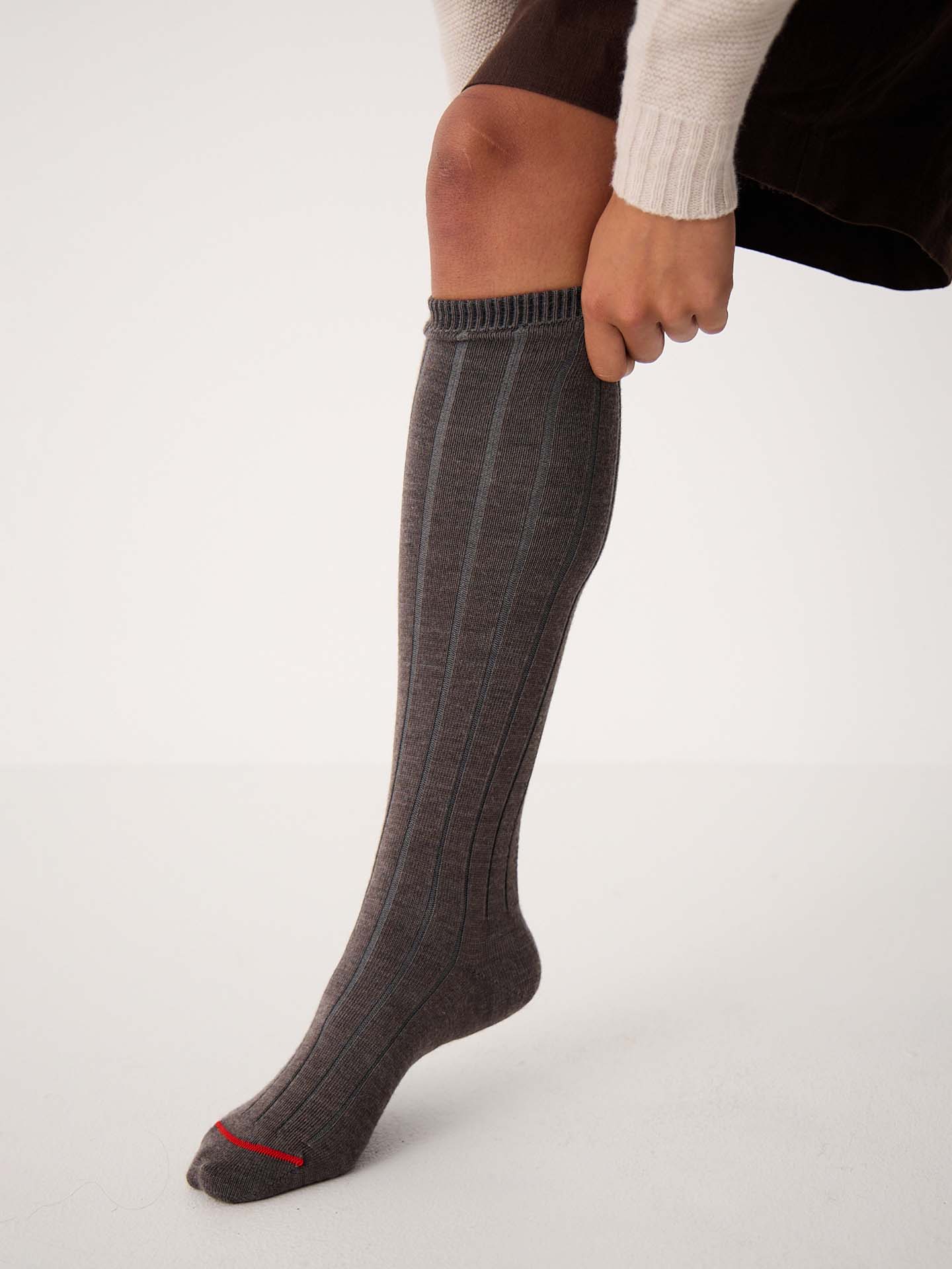 Wearing Tip Toe Knee High Socks in Grey with Cherry Trim, a person in a brown skirt and cream sweater pulls up the ribbed socks featuring a subtle red accent near the toe.