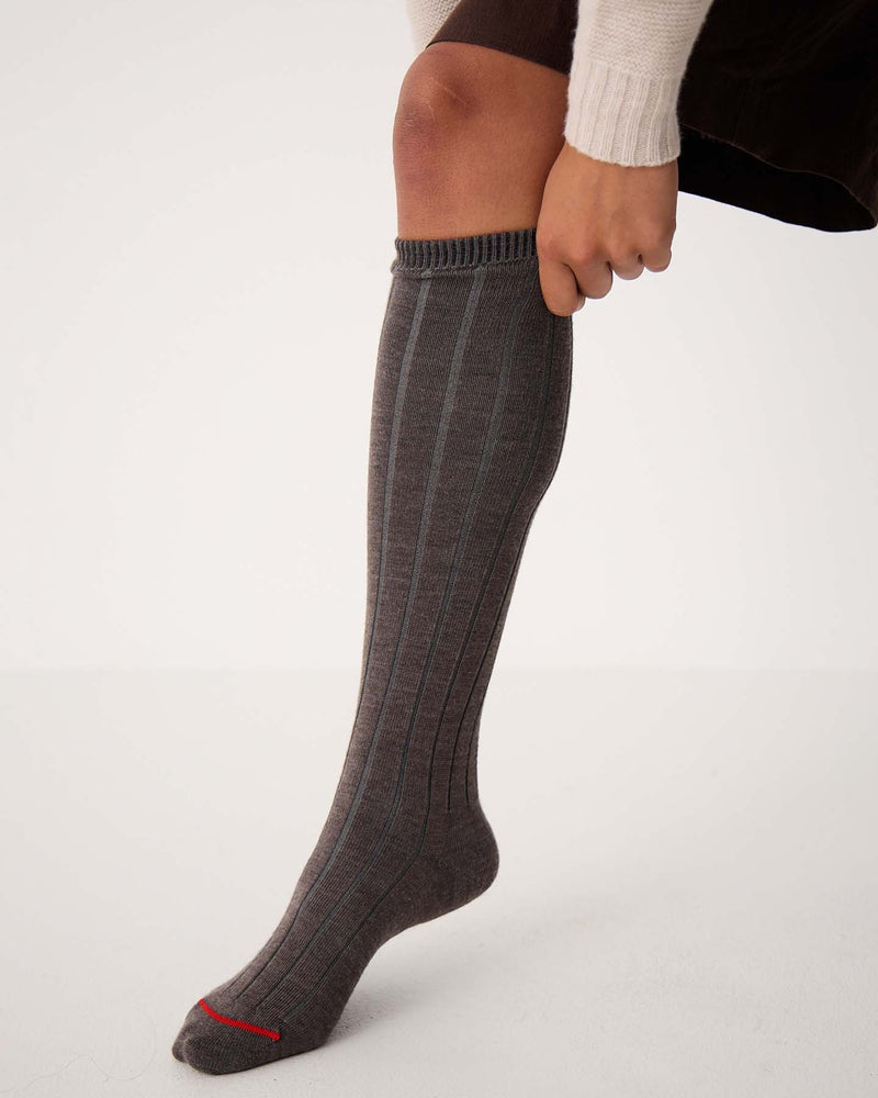 Wearing Tip Toe Knee High Socks in Grey with Cherry Trim, a person in a brown skirt and cream sweater pulls up the ribbed socks featuring a subtle red accent near the toe.