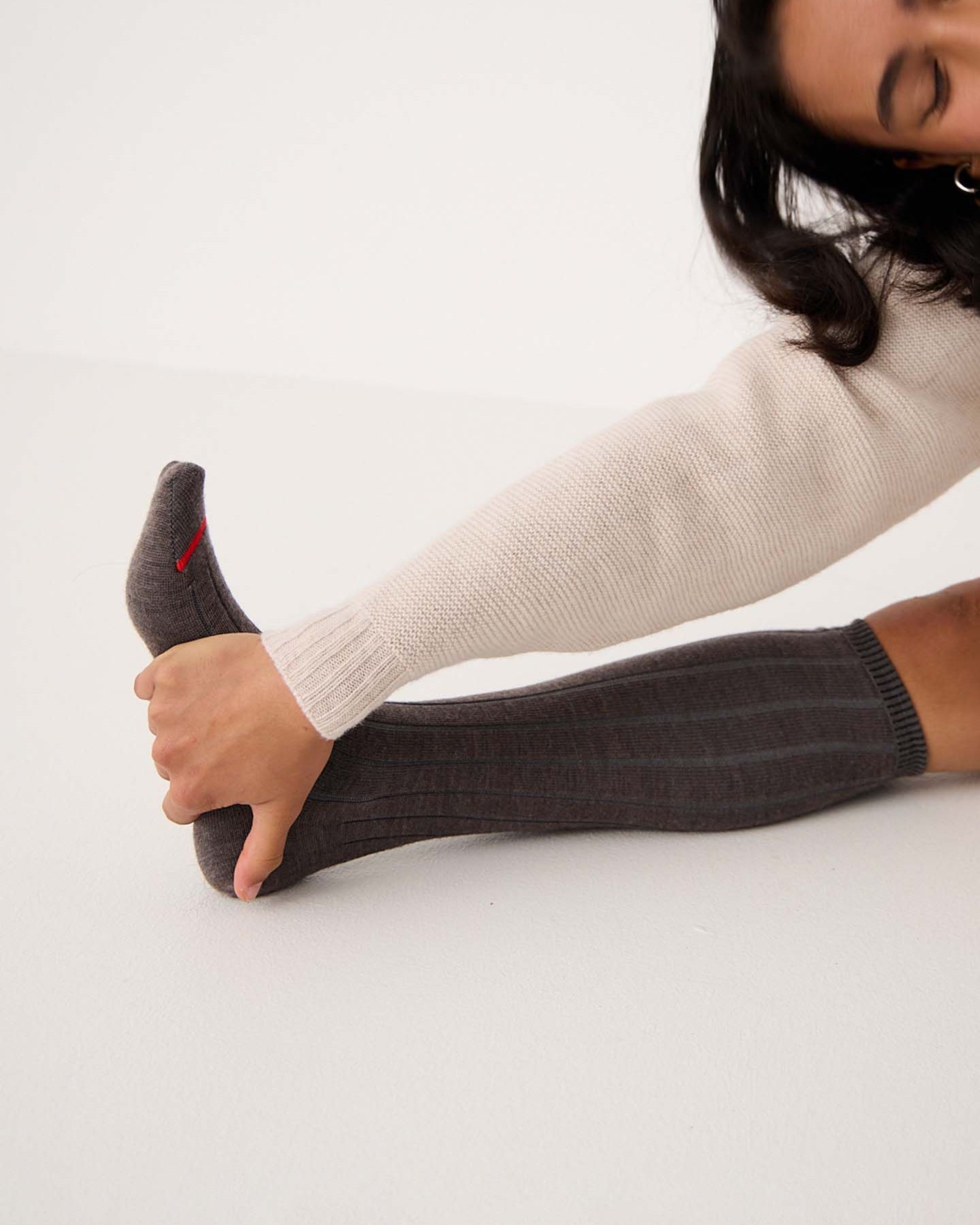 Wearing Tip Toe Knee High Socks in Grey with Cherry Trim, a person in a brown skirt and cream sweater pulls up the ribbed socks featuring a subtle red accent near the toe.