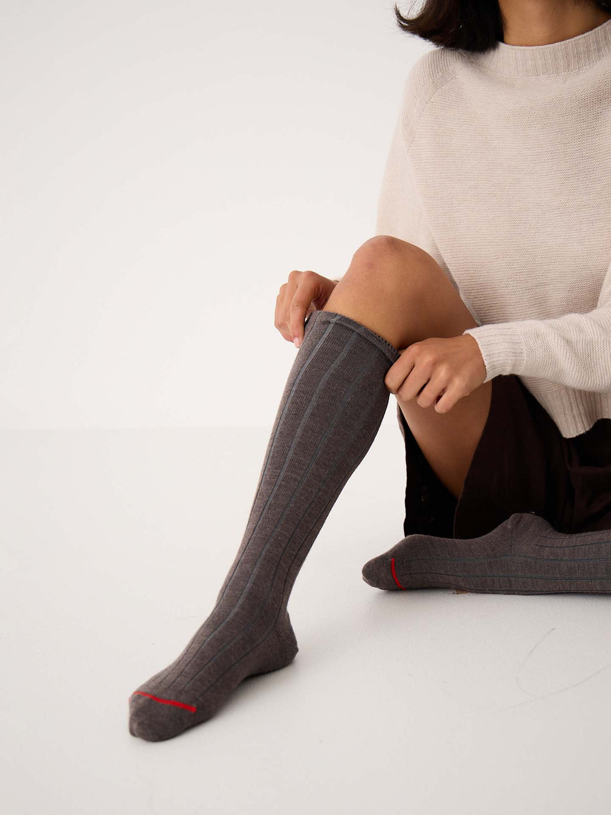 A person sits on the floor putting on Tip Toe Knee High Socks – Grey & Cherry Trim, available in small, medium, and large sizes, while wearing a light sweater and a dark skirt.