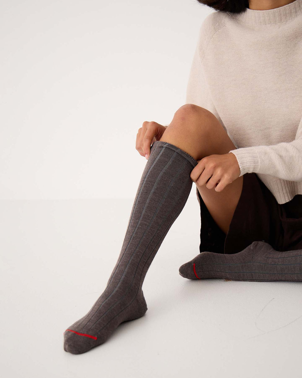 A person sits on the floor putting on Tip Toe Knee High Socks – Grey & Cherry Trim, available in small, medium, and large sizes, while wearing a light sweater and a dark skirt.