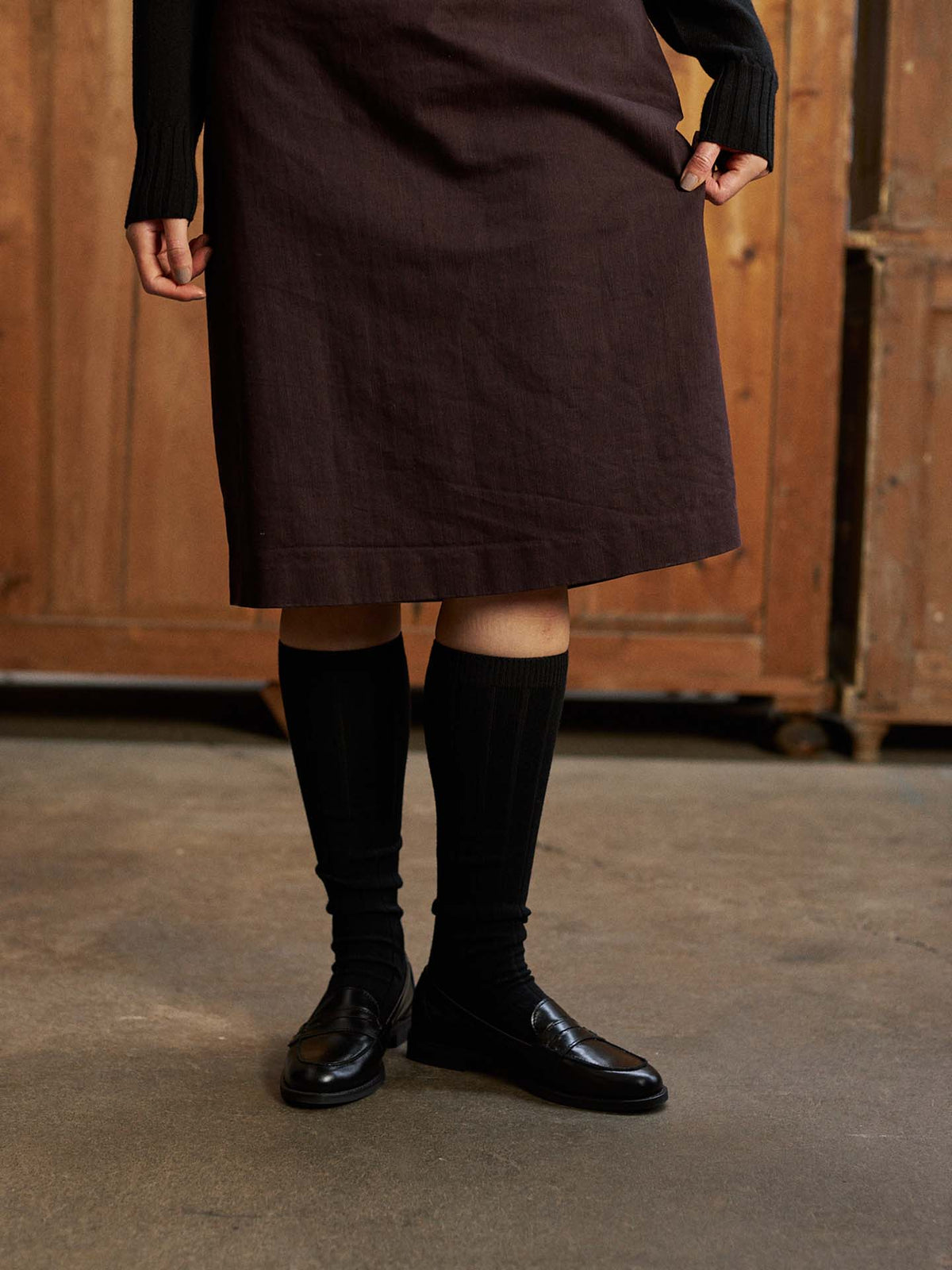 Someone indoors models the Tip Toe Knee High Socks – Black & Chestnut Trim with a dark skirt and black loafers—ideal for using a shoe size chart to pick the best EU, UK, or US fit; upper body mostly out of frame.