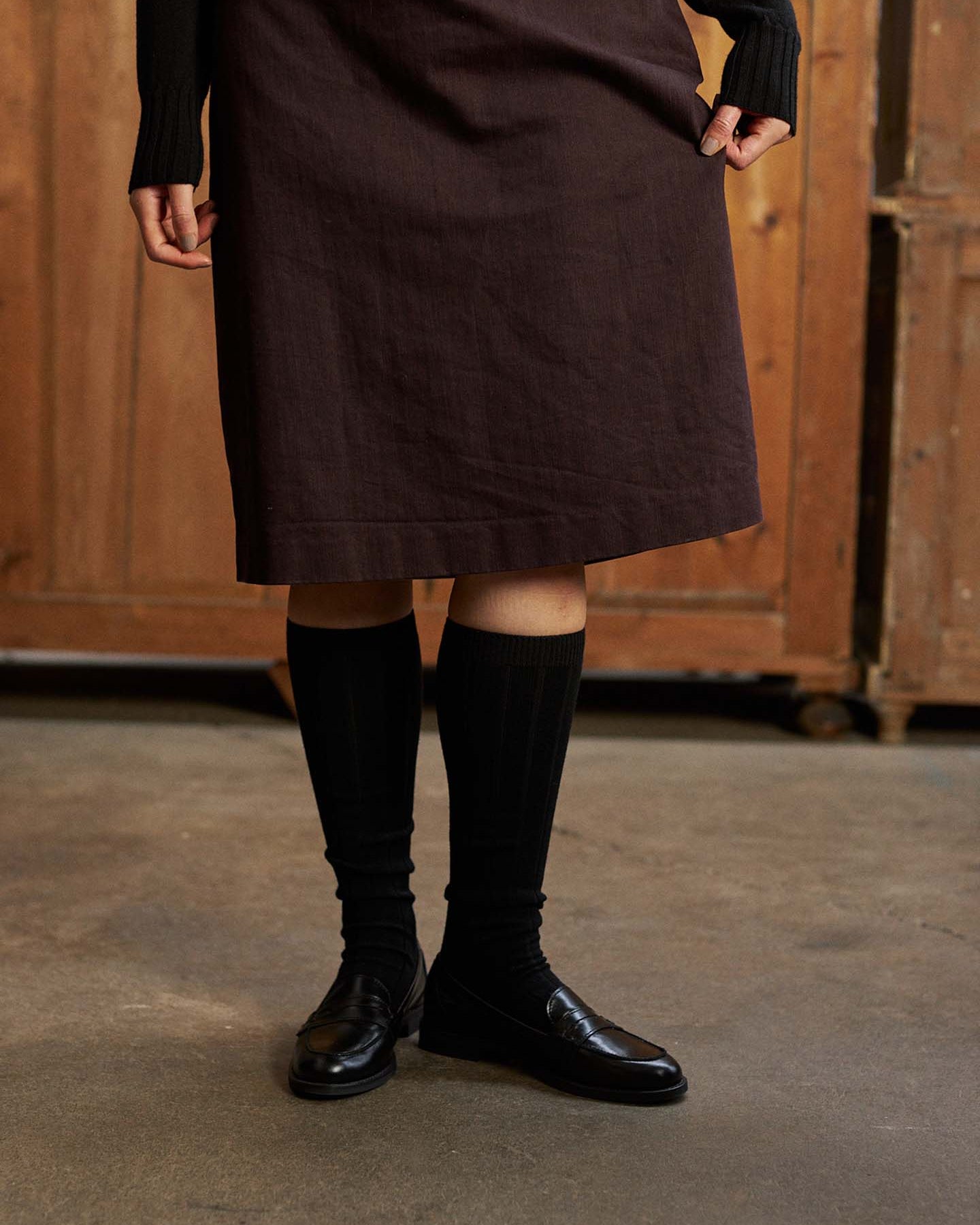 Someone indoors models the Tip Toe Knee High Socks – Black & Chestnut Trim with a dark skirt and black loafers—ideal for using a shoe size chart to pick the best EU, UK, or US fit; upper body mostly out of frame.