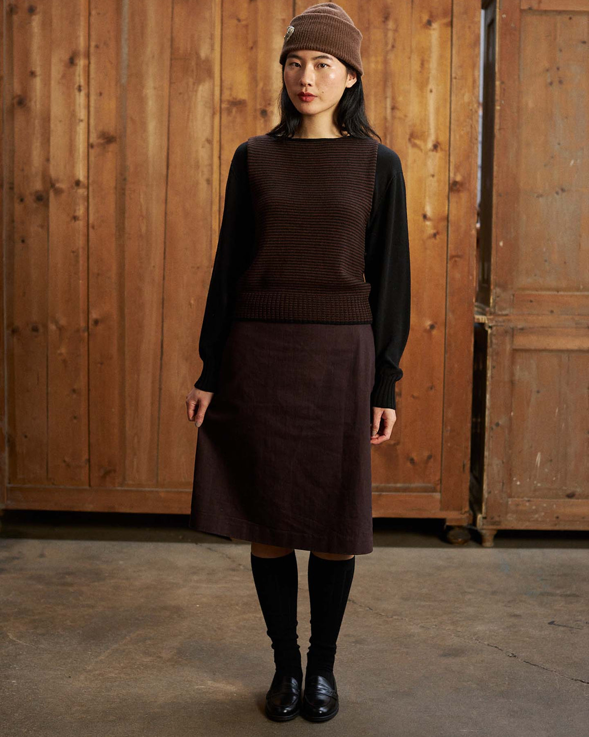 A woman stands indoors in front of wooden panels wearing Tip Toe Knee High Socks – Black & Chestnut Trim—check our size guide for EU, UK, and US sizes to find your perfect fit.