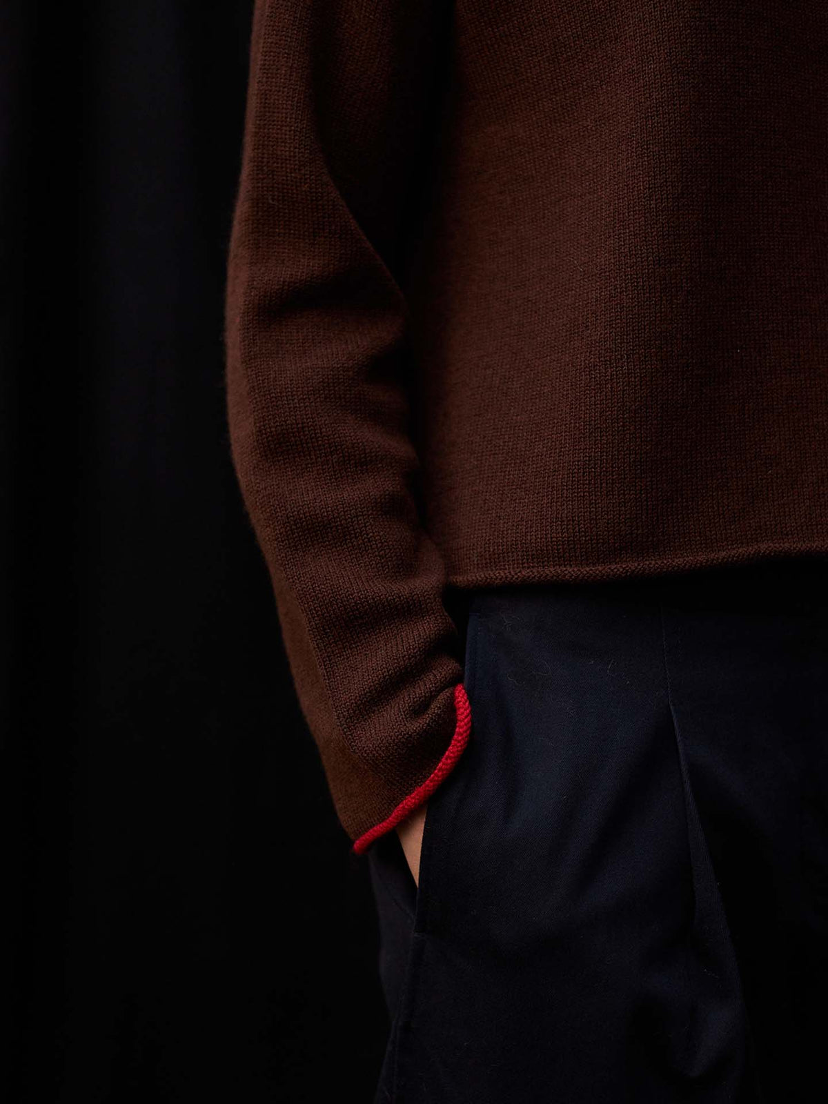 A petite person wears the Trimmed Doll's Cardigan – Chestnut & Cherry, featuring brown with red cuff detail, paired with relaxed-fit dark pants, standing against a black background with one hand in their pocket.