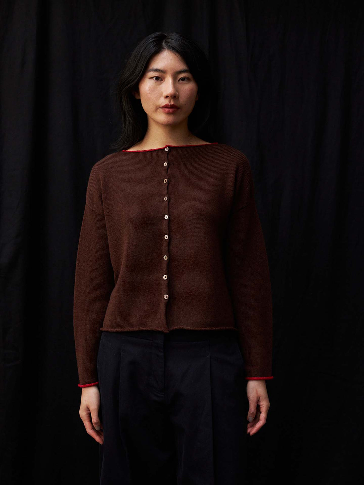 A petite woman stands before a black background, facing the camera in the Trimmed Doll's Cardigan – Chestnut & Cherry, a relaxed-fit brown sweater with red trim, paired with black pants and a neutral expression.