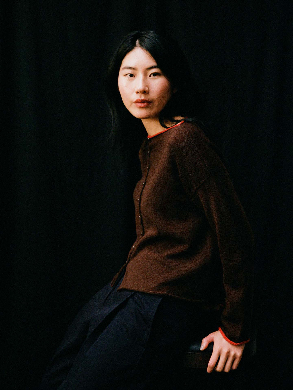 A petite person with long black hair sits on a stool against a dark background, wearing the Trimmed Doll's Cardigan – Chestnut & Cherry and dark pants.
