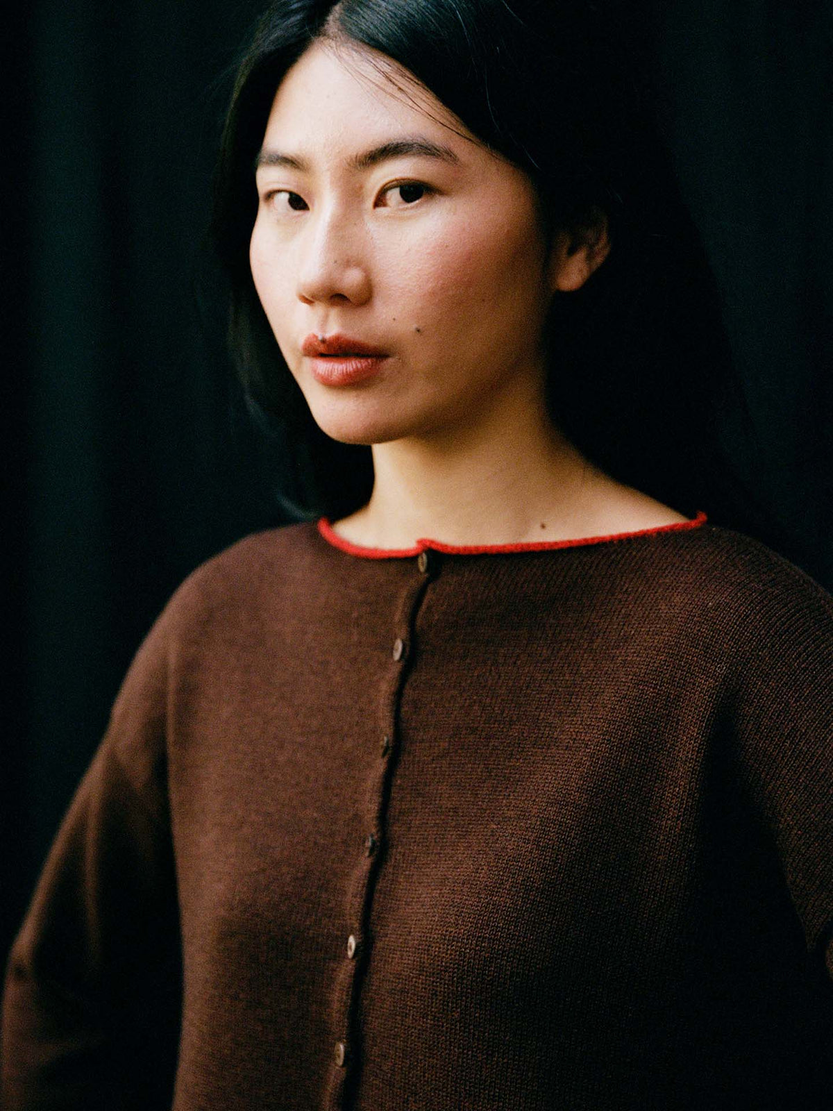 A petite woman with long black hair wears the Trimmed Doll's Cardigan in Chestnut & Cherry—a brown, relaxed-fit button-up with a red-trimmed neckline—standing before a dark background.