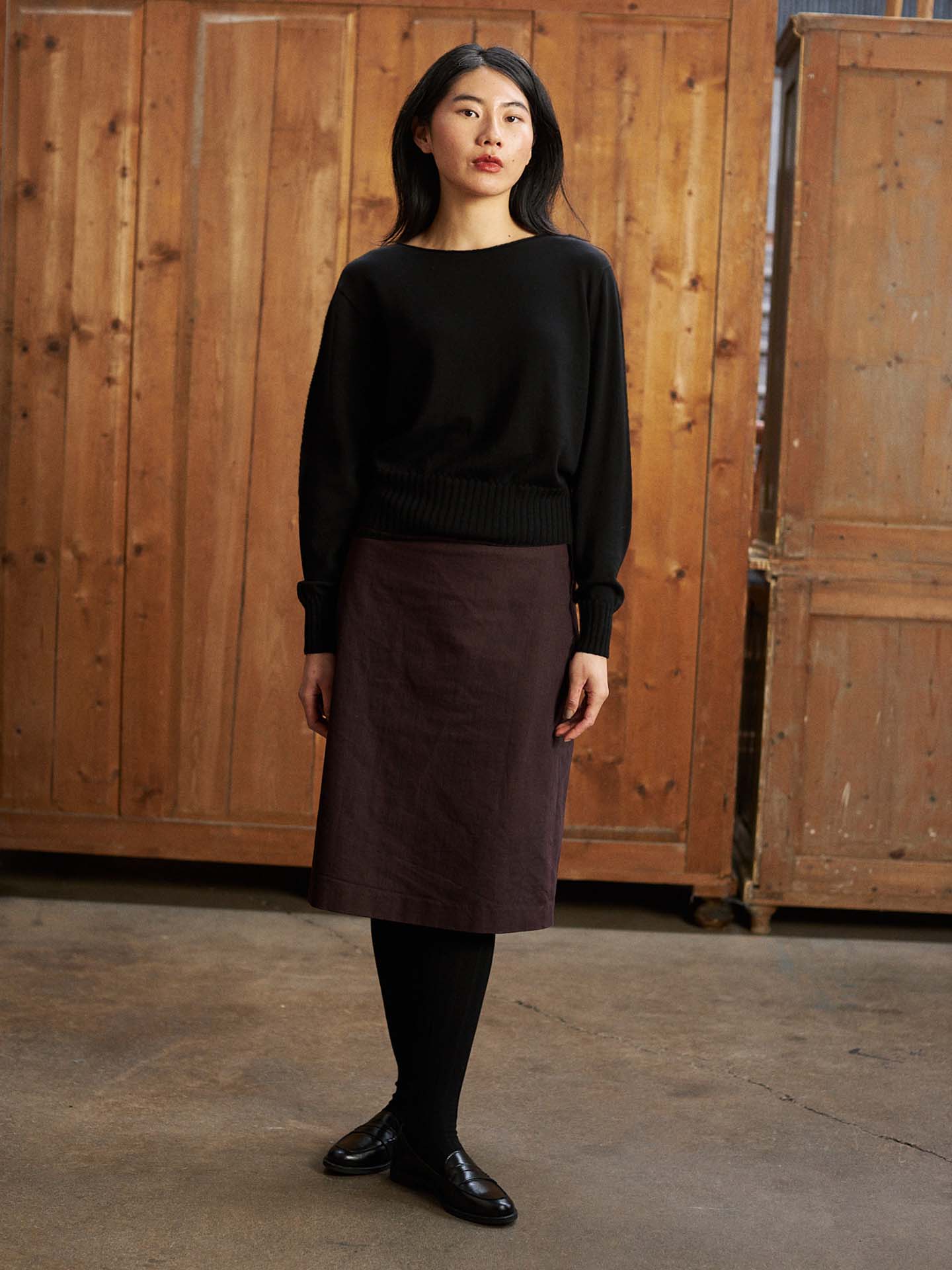 A woman with a petite fit stands indoors on a concrete floor, wearing the Trinket Knit – Black sweater, a dark skirt, black tights, and black shoes against a wooden paneled background.