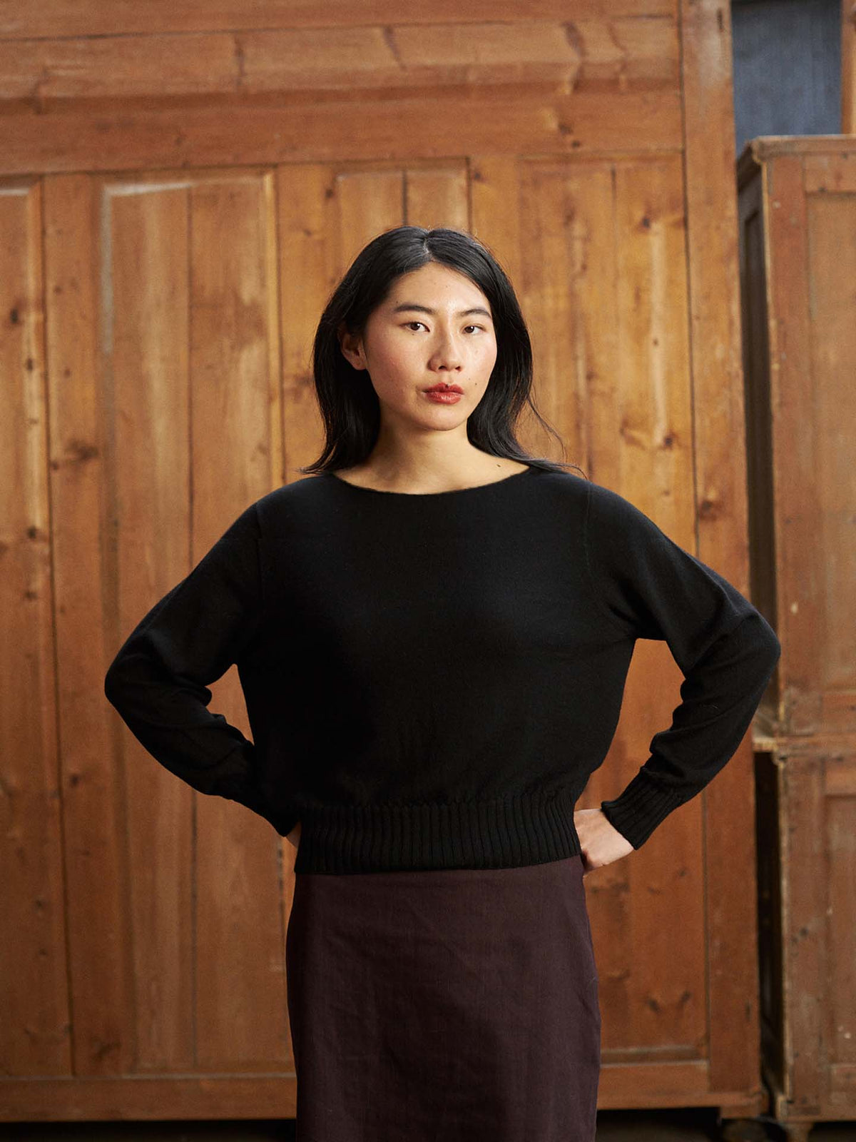 A woman with long dark hair poses indoors in front of wooden paneled doors, wearing the Trinket Knit – Black sweater in a relaxed fit and a dark skirt, her hands resting on her hips.