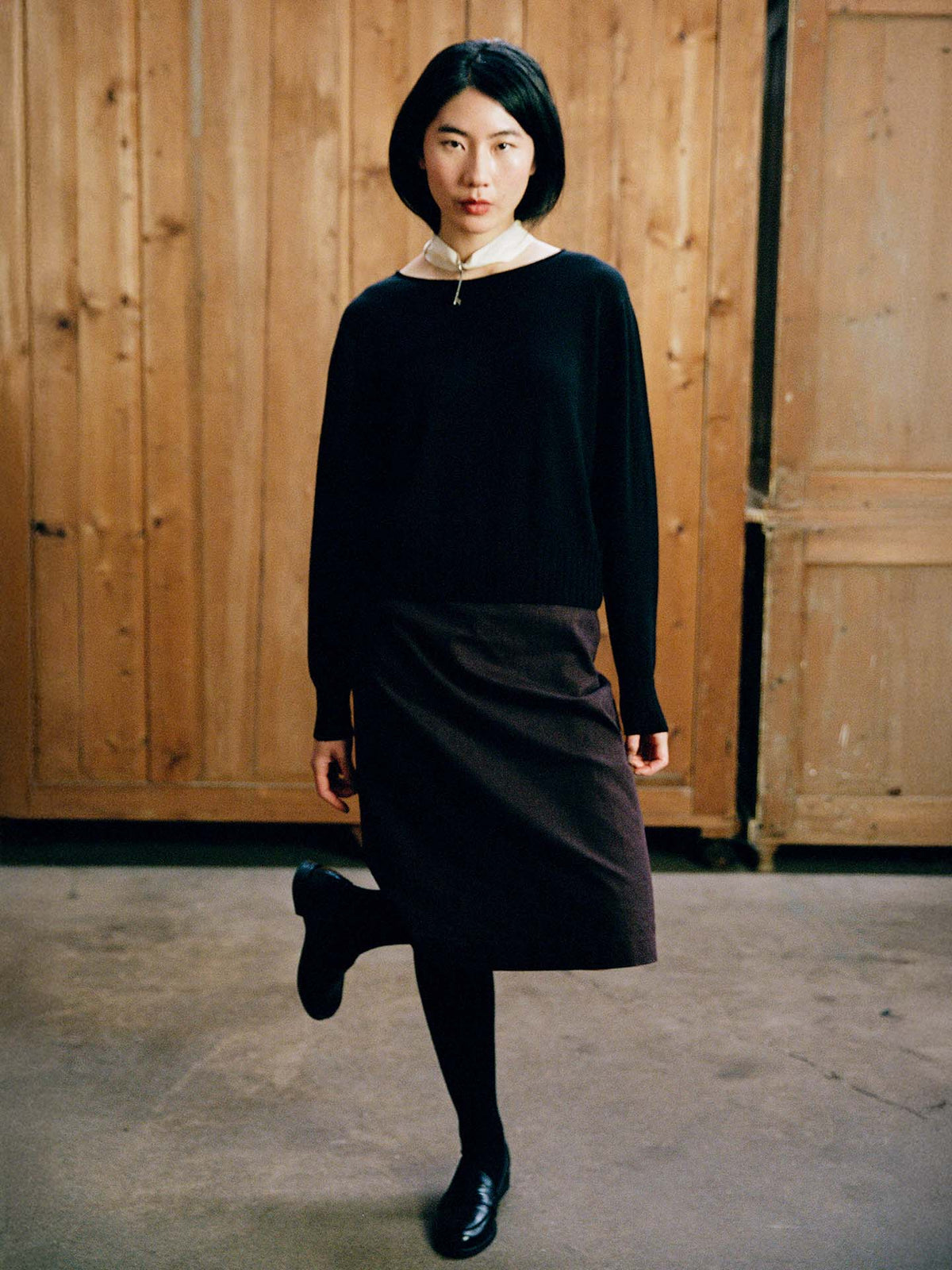 A person with straight black hair stands indoors on one leg in front of wooden paneled doors, wearing the Trinket Knit – Black in a petite fit with a white collar.