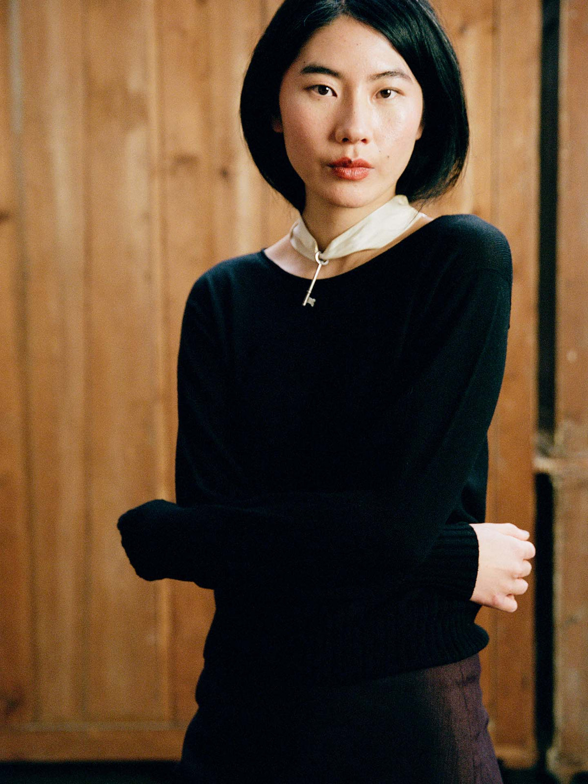 A petite woman with short dark hair, wearing the Trinket Knit – Black sweater, stands with arms crossed in front of a wooden wall.