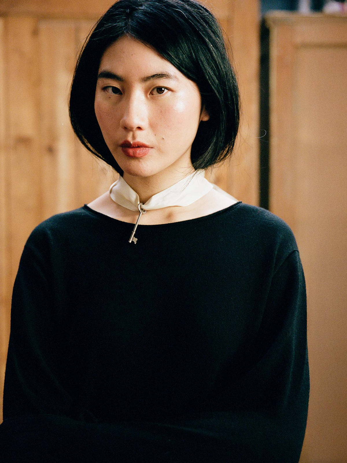 A person with straight black hair and a serious expression wears the Trinket Knit – Black, a cropped top, along with a white scarf fastened by a metallic pin, standing indoors before wooden panels.