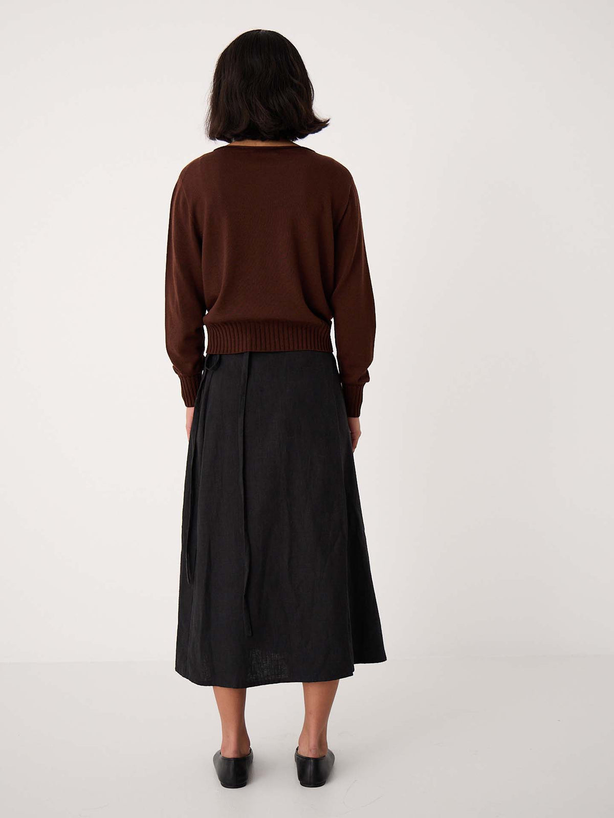 A person faces away wearing the Trinket Knit – Chestnut, a relaxed-fit cropped brown sweater, styled with a long black skirt and black shoes against a plain white background.