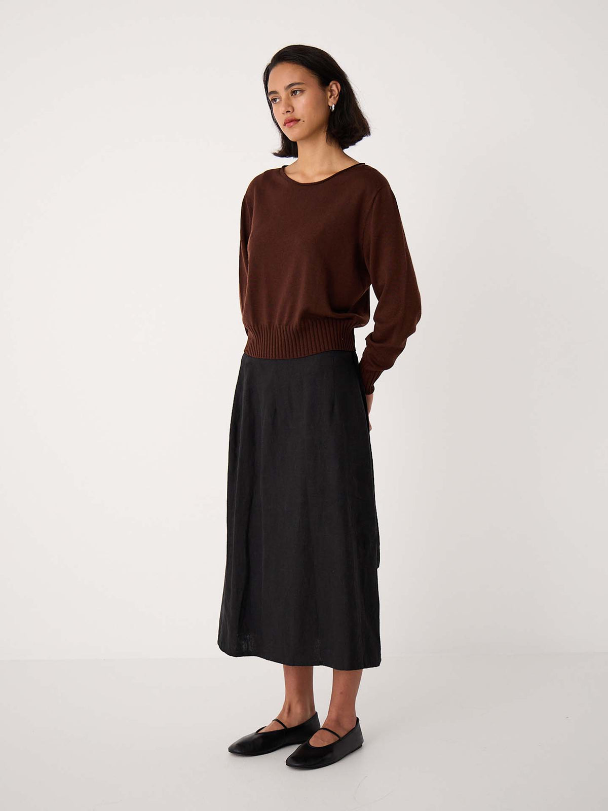 A petite woman stands in a plain studio wearing the Trinket Knit – Chestnut, a relaxed-fit brown sweater, paired with a black midi skirt and black flat shoes, looking slightly to the side.