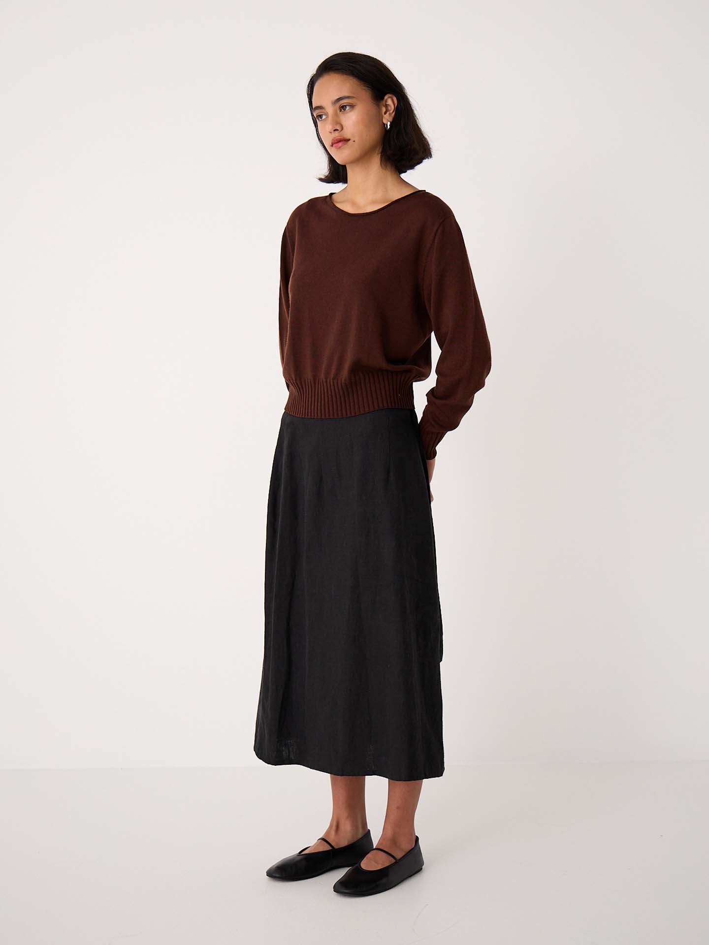 A petite woman stands in a plain studio wearing the Trinket Knit – Chestnut, a relaxed-fit brown sweater, paired with a black midi skirt and black flat shoes, looking slightly to the side.