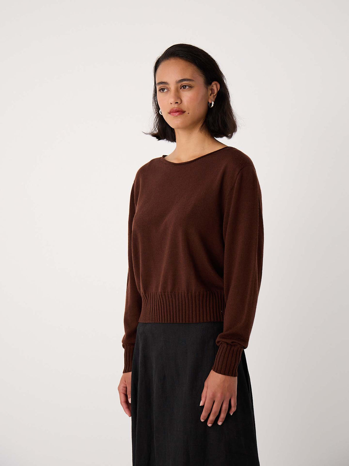 A petite woman with straight dark hair wears the Trinket Knit – Chestnut sweater and a black skirt, standing against a plain light background.