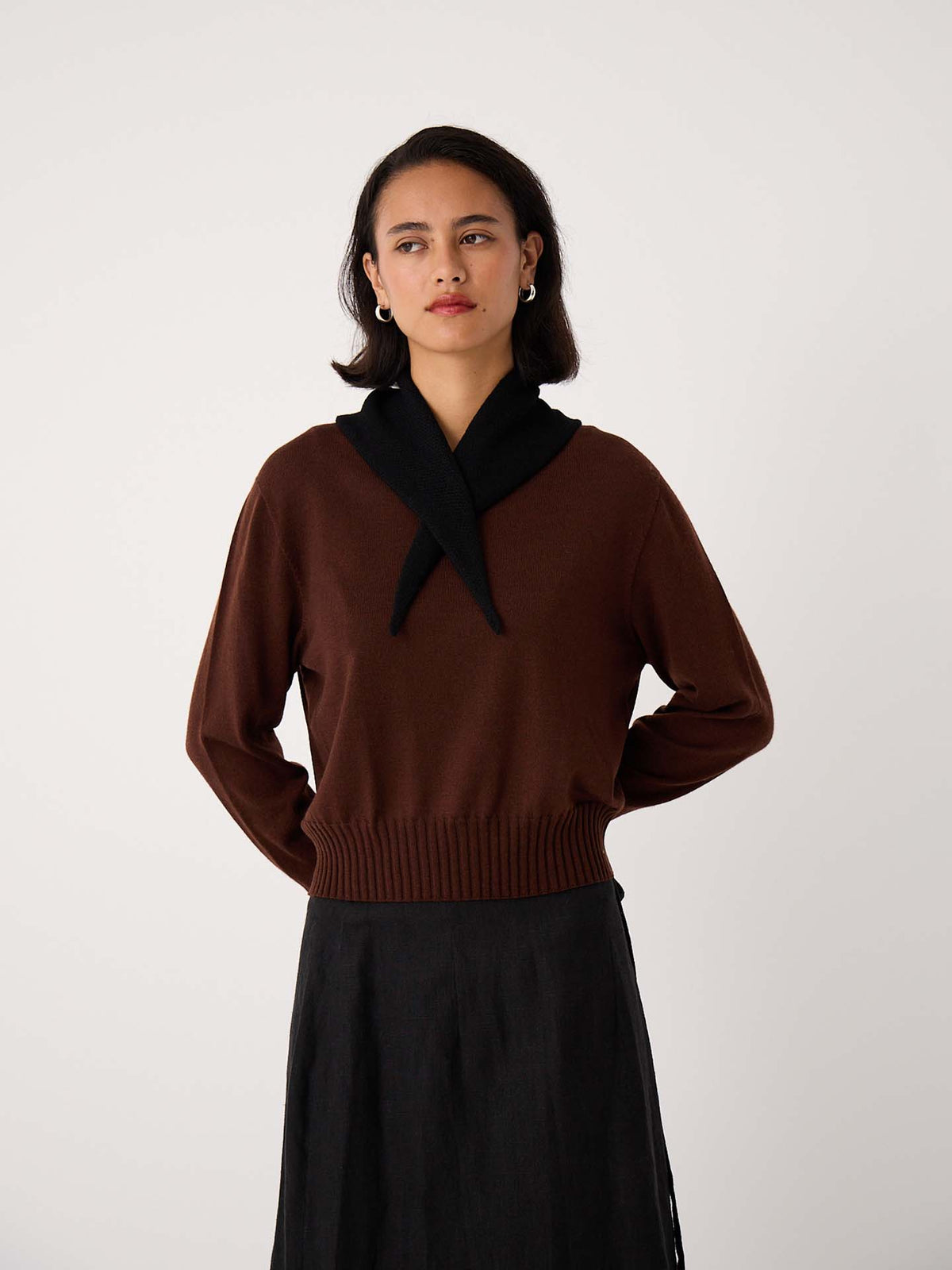 A petite woman wears the Trinket Knit – Chestnut sweater with a black sweater over her shoulders and a black skirt, standing against a plain light background.