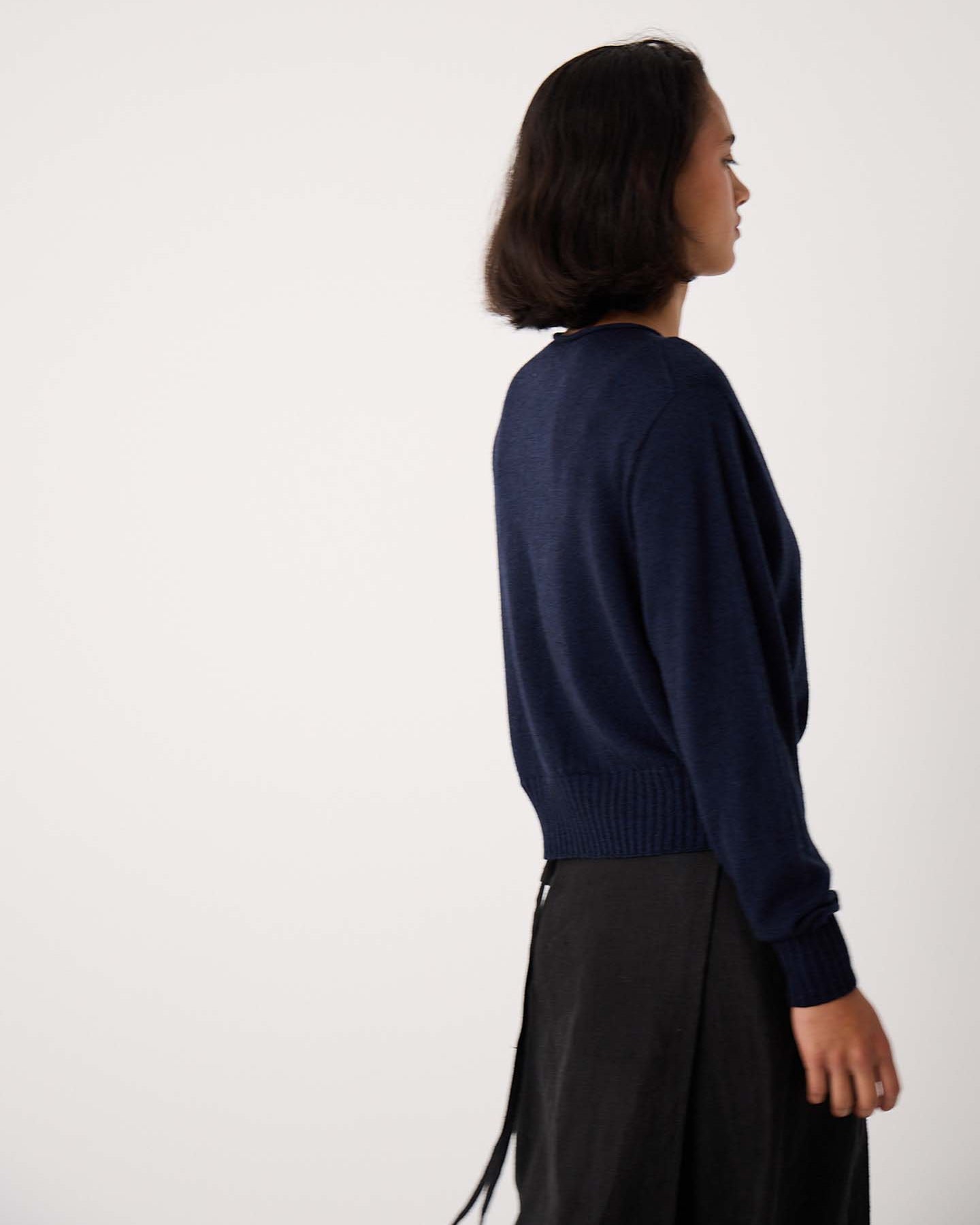 A woman stands against a plain white background wearing the Trinket Knit – Ink sweater in a relaxed fit, styled with a black midi skirt and black flats.