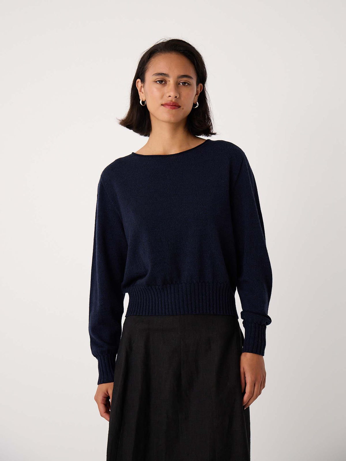 A petite woman with shoulder-length dark hair wears the Trinket Knit – Ink, a relaxed-fit navy sweater, paired with a black skirt against a plain light background.