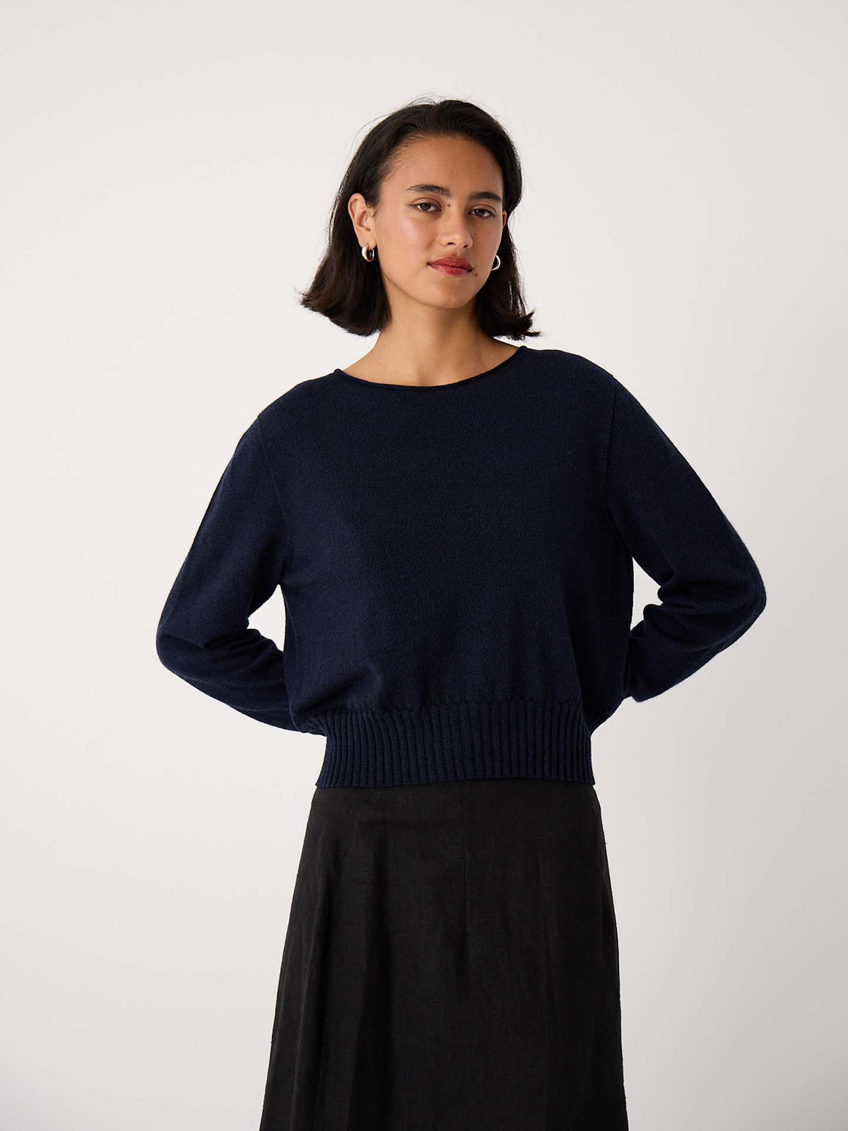 Woman with short dark hair wears the Trinket Knit – Ink, a navy blue relaxed-fit long-sleeve sweater, paired with a black skirt against a plain light background.