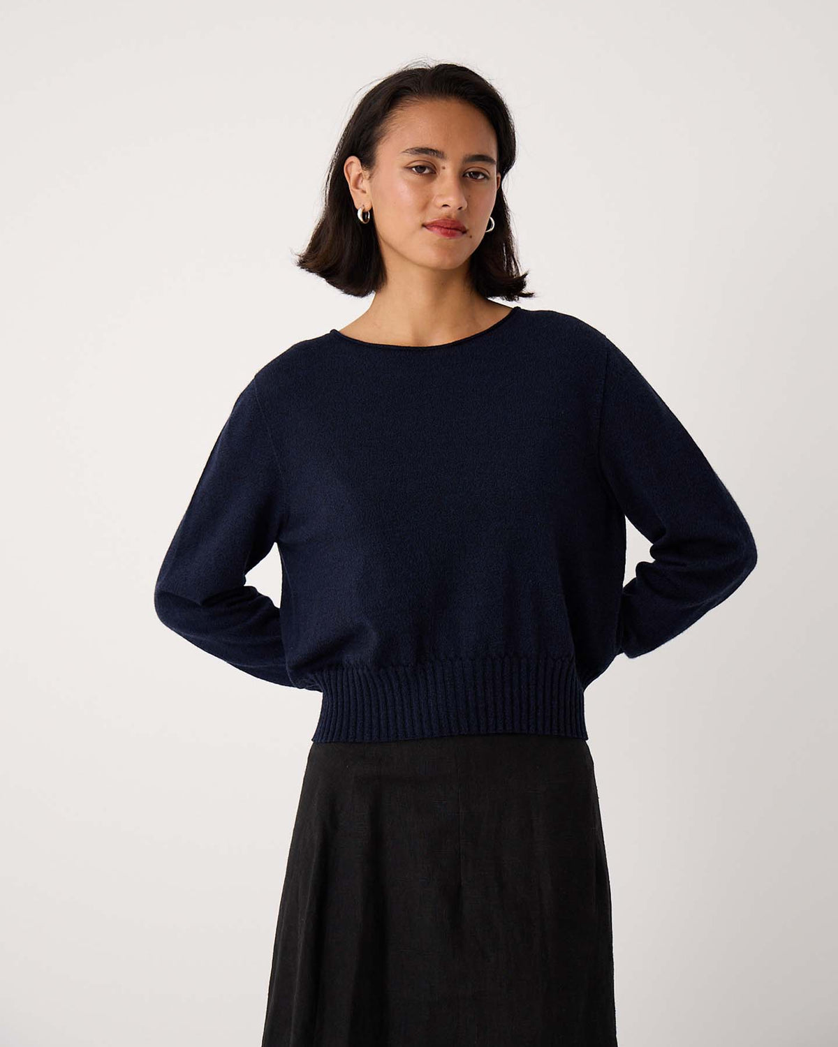 Woman with short dark hair wears the Trinket Knit – Ink, a navy blue relaxed-fit long-sleeve sweater, paired with a black skirt against a plain light background.