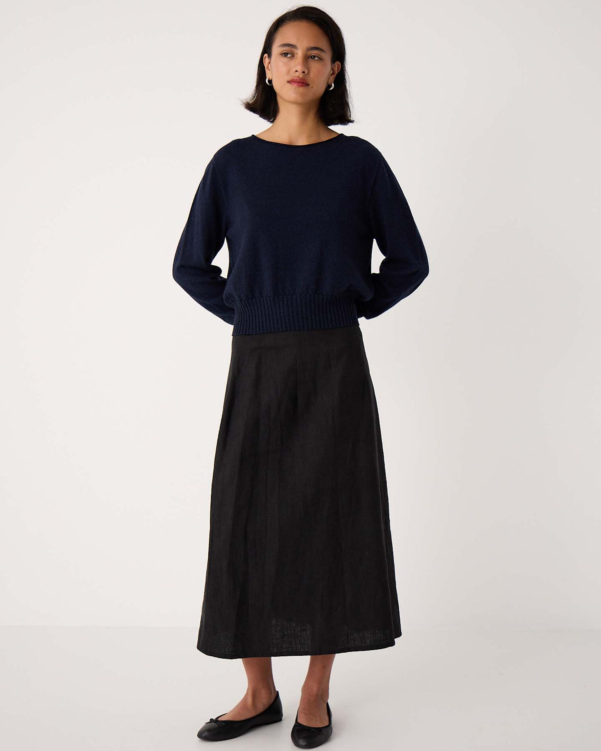 A woman stands against a plain background wearing the Trinket Knit – Ink, a relaxed-fit navy sweater, paired with a long black skirt and black flats, hands behind her back.