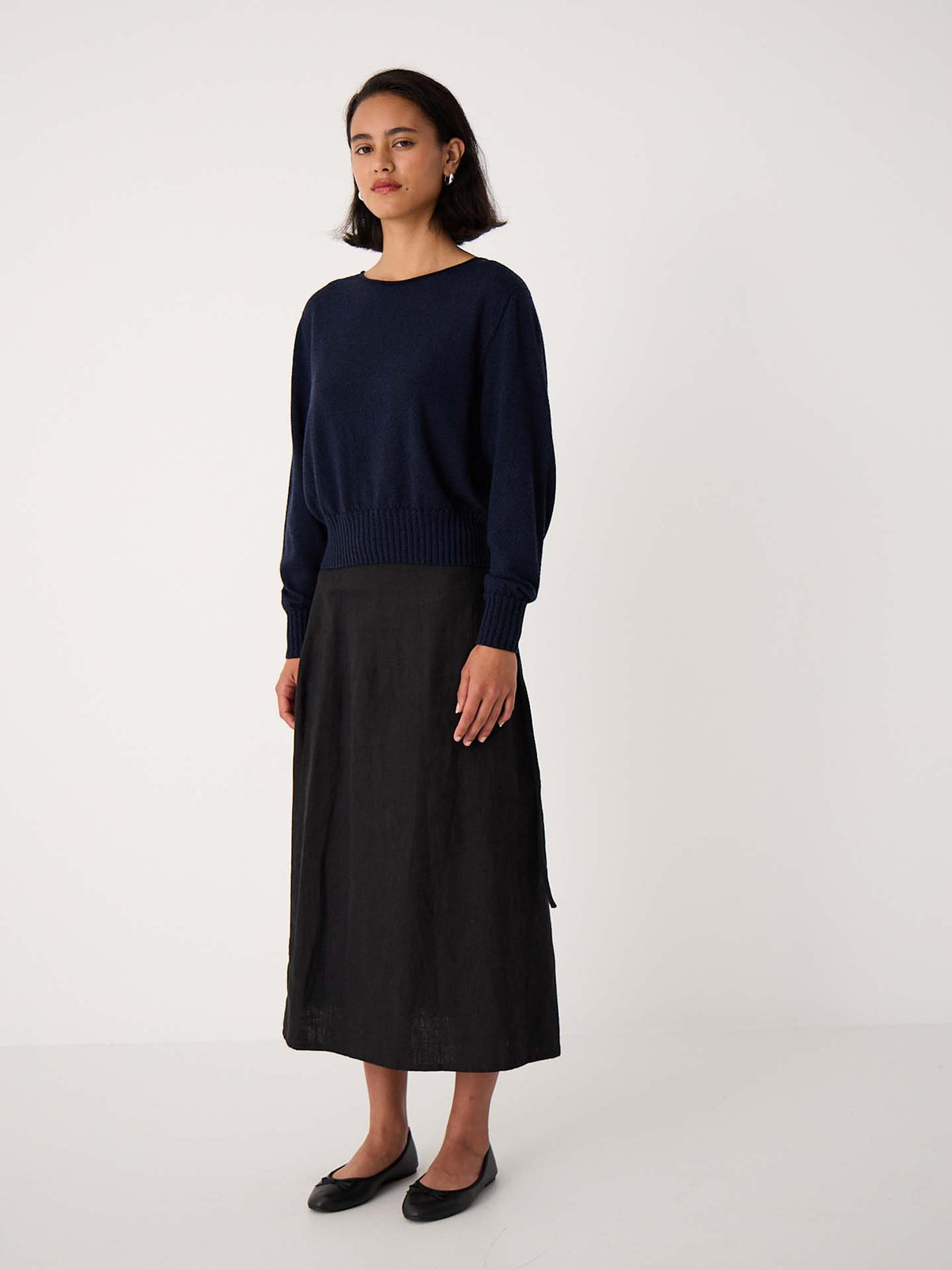 A woman stands against a plain white background wearing the Trinket Knit – Ink sweater in a relaxed fit, styled with a black midi skirt and black flats.