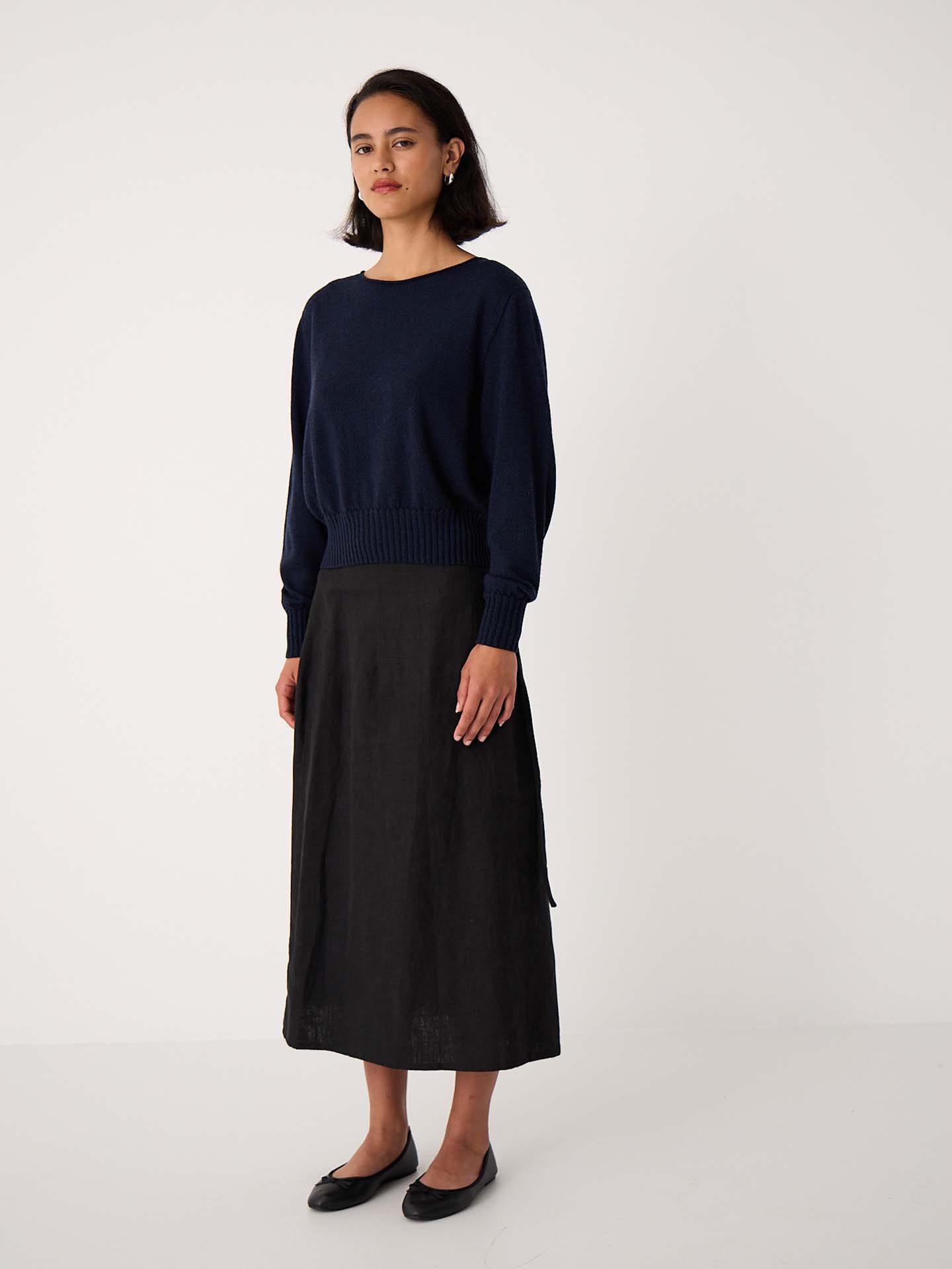 A woman stands against a plain white background wearing the Trinket Knit – Ink sweater in a relaxed fit, styled with a black midi skirt and black flats.