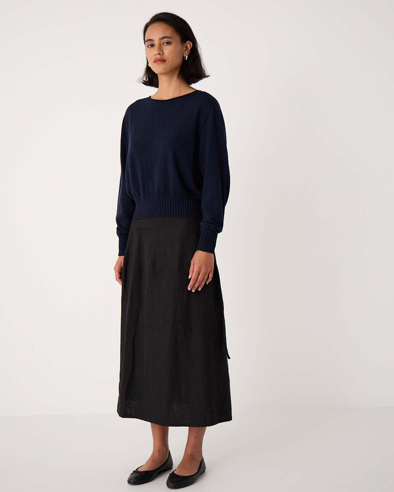 A woman stands against a plain white background wearing the Trinket Knit – Ink sweater in a relaxed fit, styled with a black midi skirt and black flats.