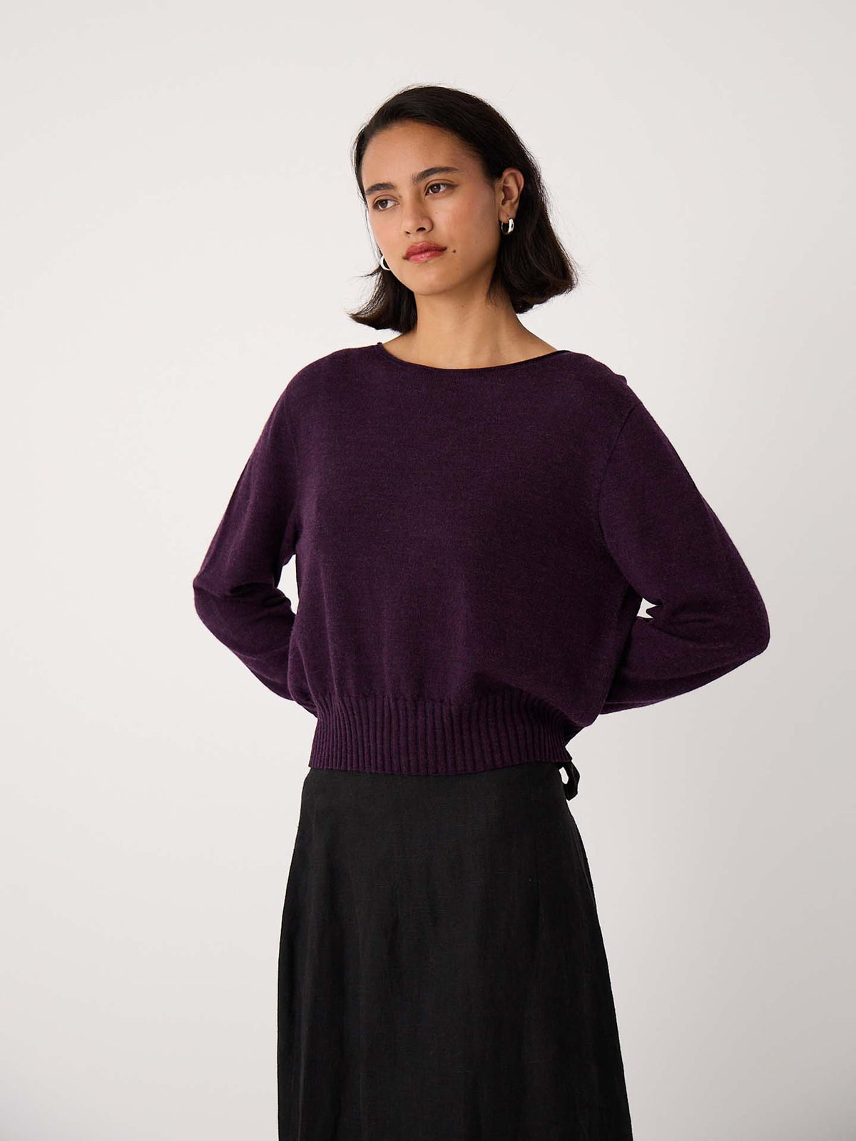 A woman stands against a plain background wearing the Trinket Knit – Malbec, a dark purple long-sleeve relaxed fit sweater, paired with a black skirt and her hands on her hips.