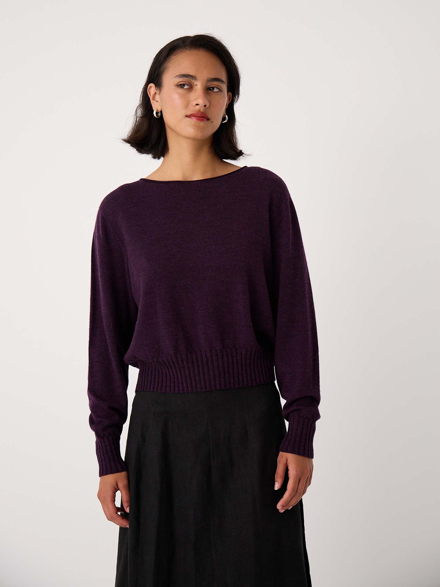 A woman stands against a plain background, wearing the Trinket Knit – Malbec, a dark purple relaxed fit long-sleeve sweater, paired with a black skirt.