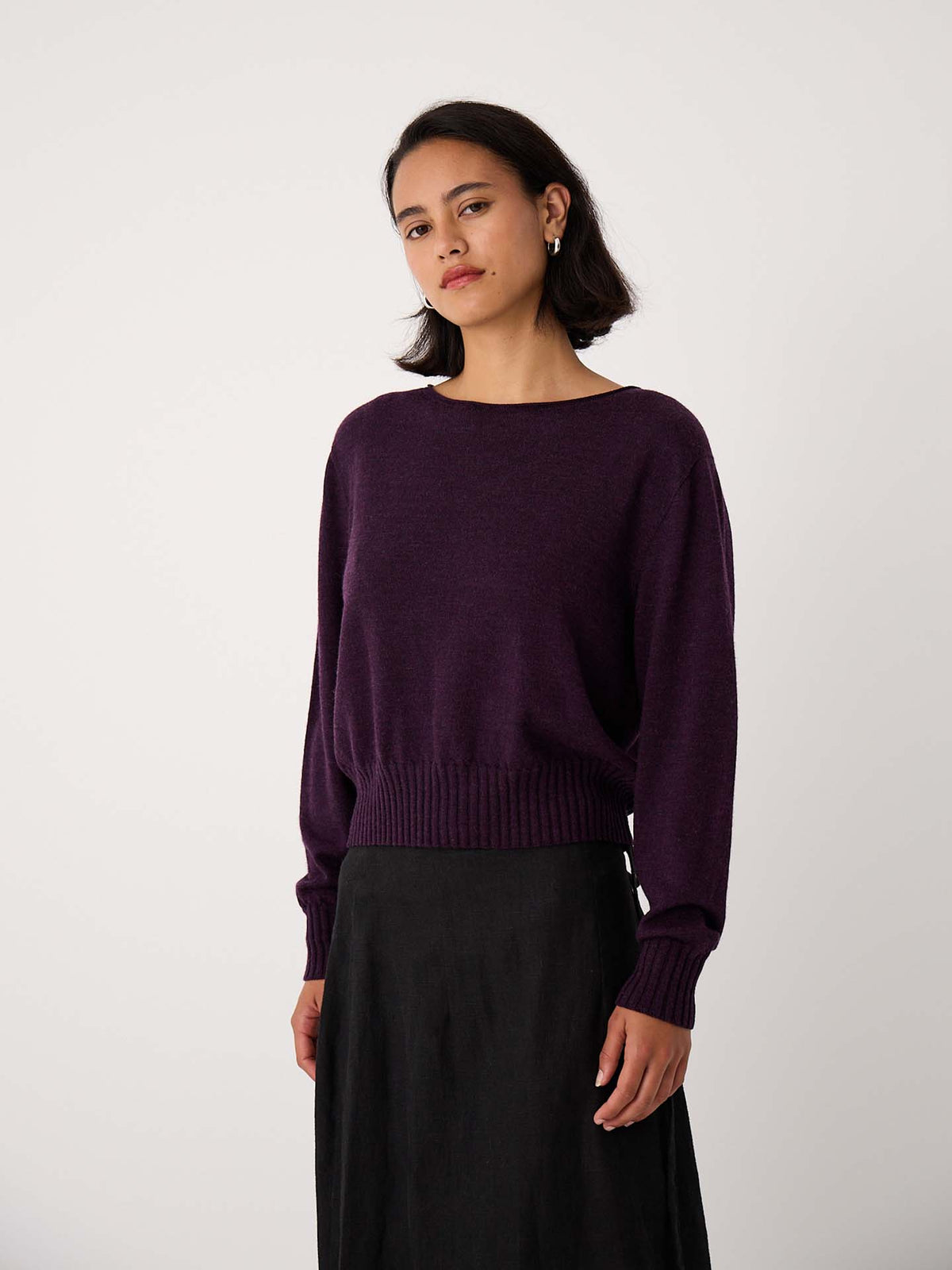 A woman with shoulder-length dark hair wears the Trinket Knit – Malbec, a purple relaxed fit sweater, paired with a black skirt and stands against a plain light background.