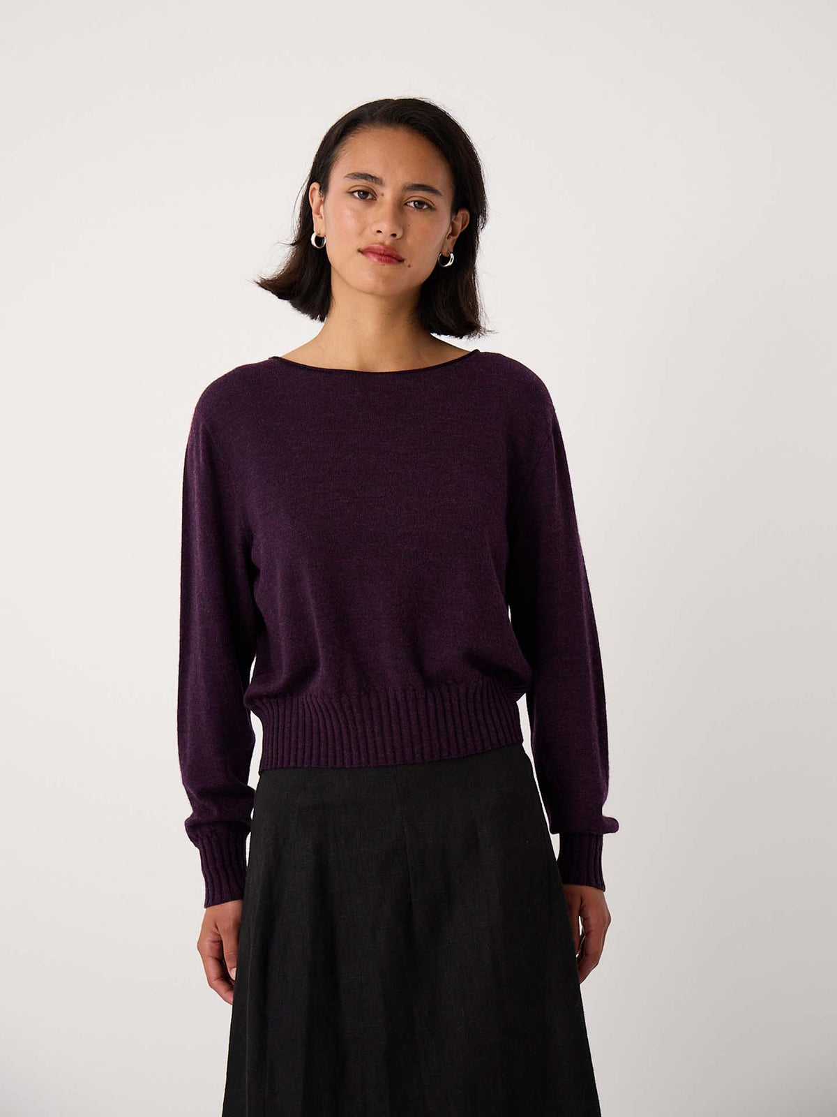 A woman stands against a plain background, wearing the Trinket Knit – Malbec, a dark purple relaxed fit long-sleeve sweater paired with a black skirt—ideal for stylish petite fashion.