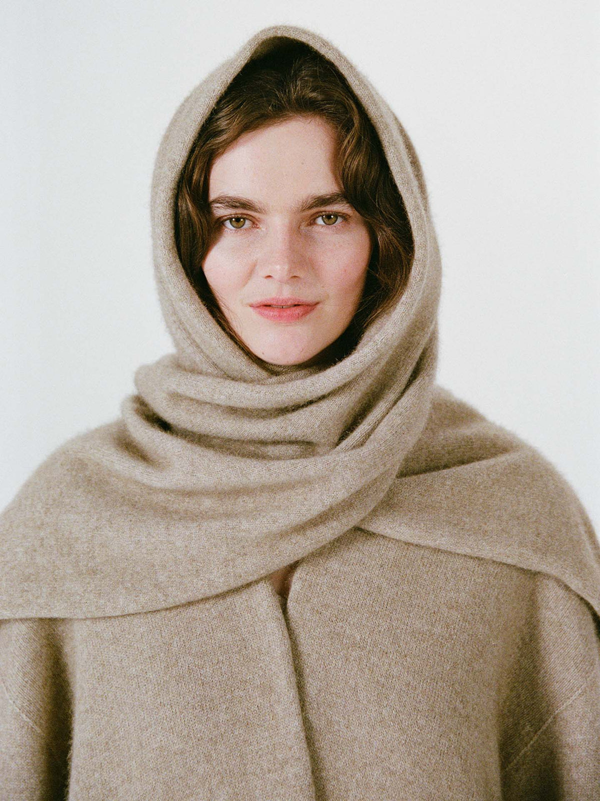 A woman wearing the Umbra Scarf – Natural, a beige wool hooded garment, stands beside an extra wide yoga mat, her serene presence highlighted against a plain light background.