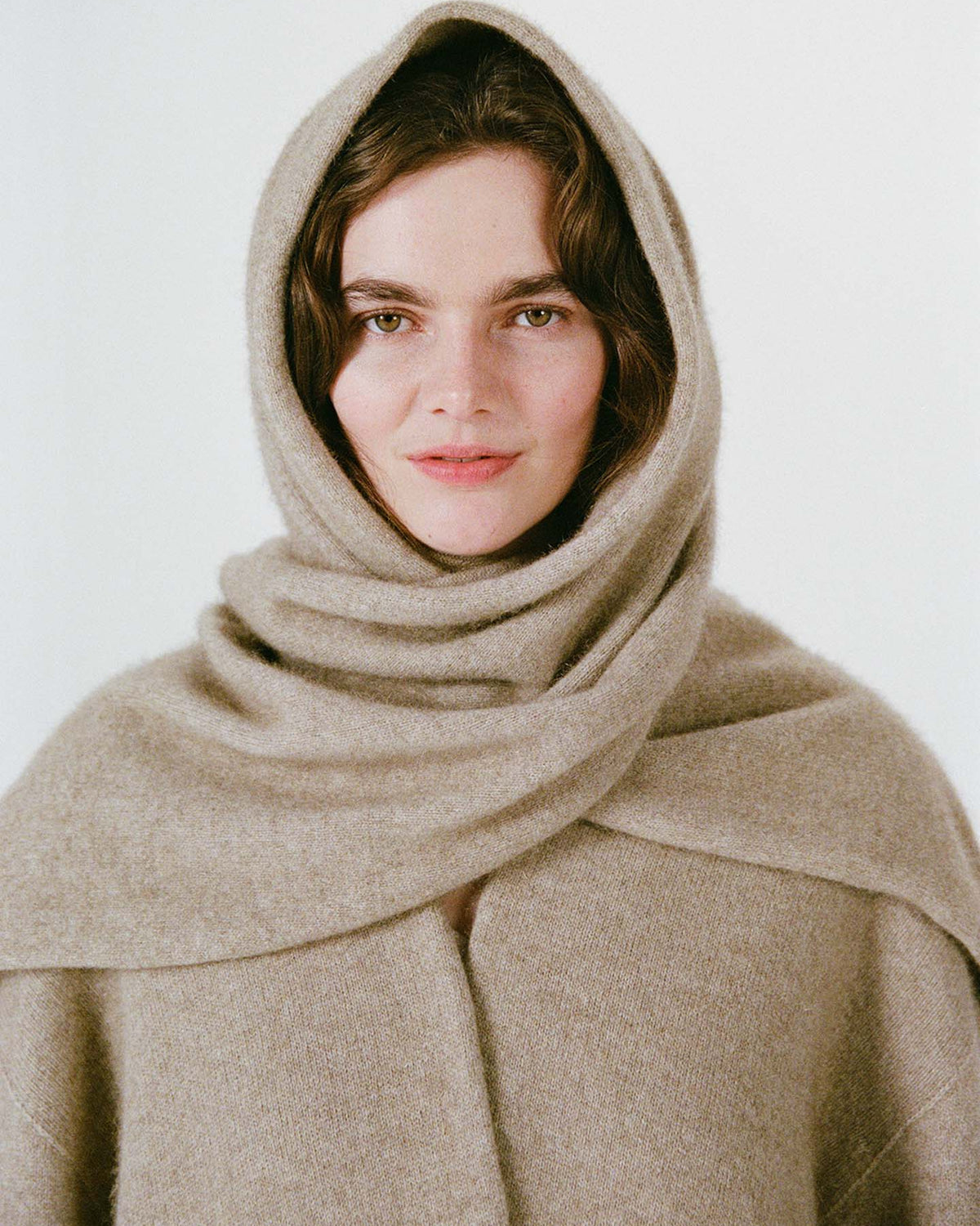 A woman wearing the Umbra Scarf – Natural, a beige wool hooded garment, stands beside an extra wide yoga mat, her serene presence highlighted against a plain light background.