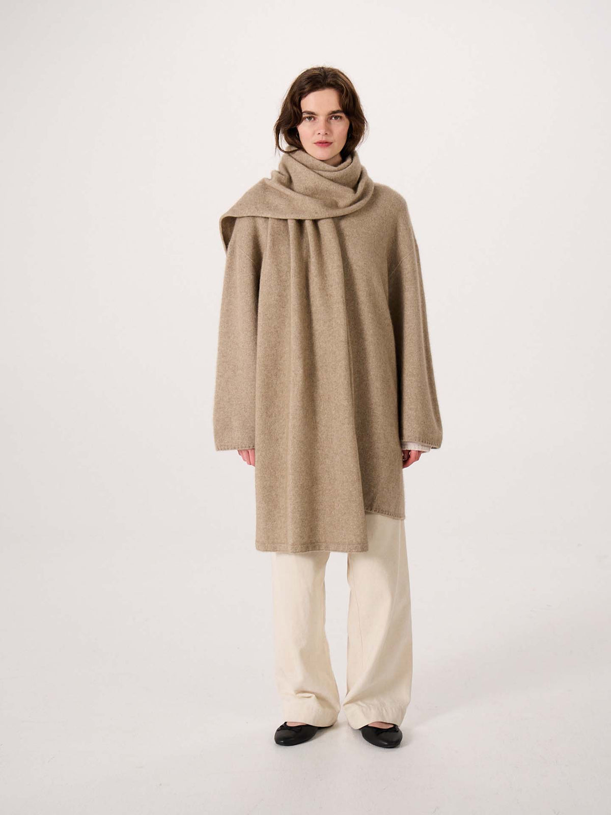 A person stands confidently on an extra wide yoga mat against a plain white background, wearing a loose camel-colored poncho paired with the Umbra Scarf – Natural, cream wide-leg pants, and black shoes.