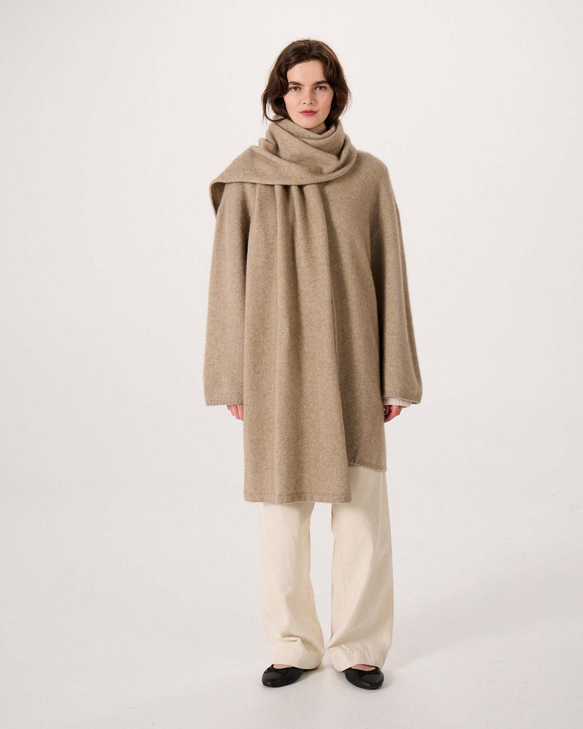 A person stands confidently on an extra wide yoga mat against a plain white background, wearing a loose camel-colored poncho paired with the Umbra Scarf – Natural, cream wide-leg pants, and black shoes.