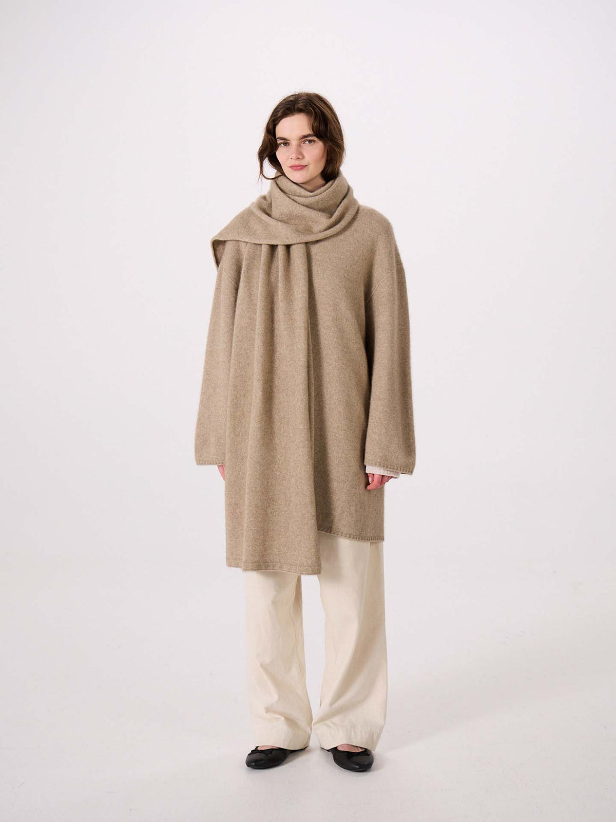 A person stands against a plain background wearing a loose beige wrap coat with the Umbra Scarf in Natural, cream pants, and black shoes, next to an extra wide yoga mat.