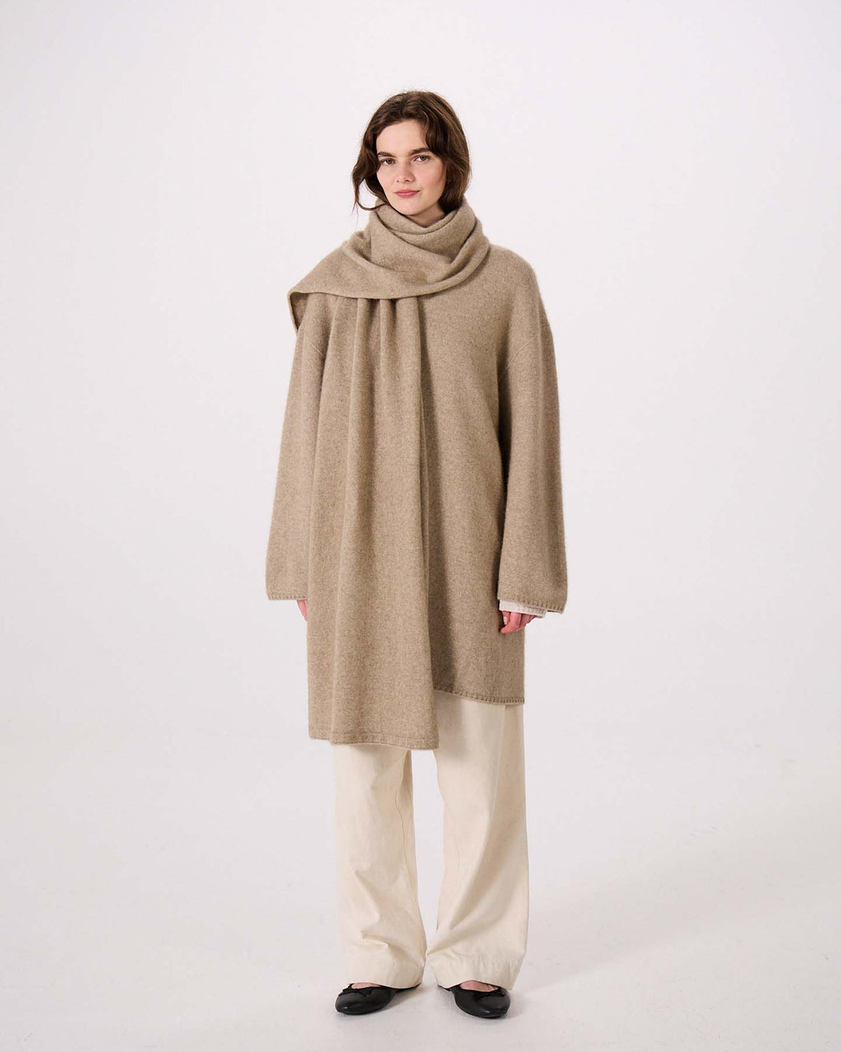 A person stands against a plain background wearing a loose beige wrap coat with the Umbra Scarf in Natural, cream pants, and black shoes, next to an extra wide yoga mat.