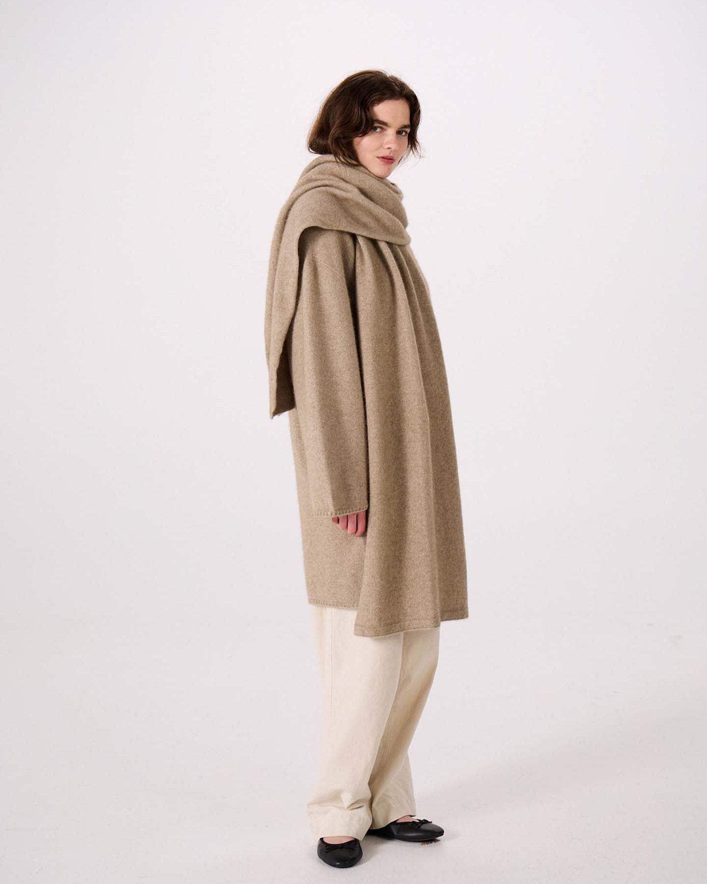Person wearing a long beige coat with the Umbra Scarf – Natural draped around their neck, paired with cream pants and black shoes, standing against a plain white background. The coat’s elegant silhouette highlights its 97cm long design, offering both style and comfort.