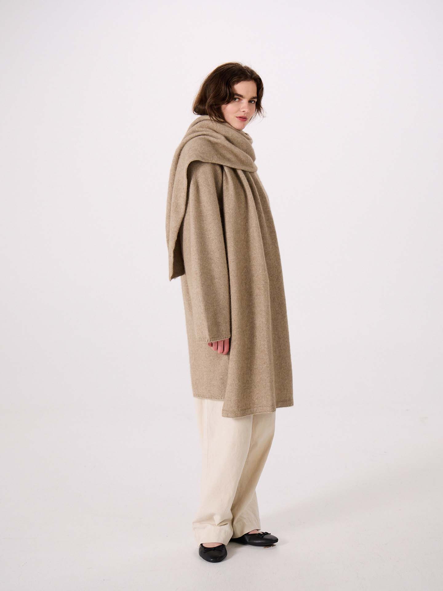 Person wearing a long beige coat with the Umbra Scarf – Natural draped around their neck, paired with cream pants and black shoes, standing against a plain white background. The coat’s elegant silhouette highlights its 97cm long design, offering both style and comfort.