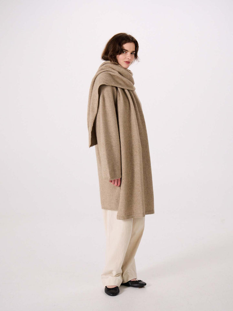 Person wearing a long beige coat with the Umbra Scarf – Natural draped around their neck, paired with cream pants and black shoes, standing against a plain white background. The coat’s elegant silhouette highlights its 97cm long design, offering both style and comfort.
