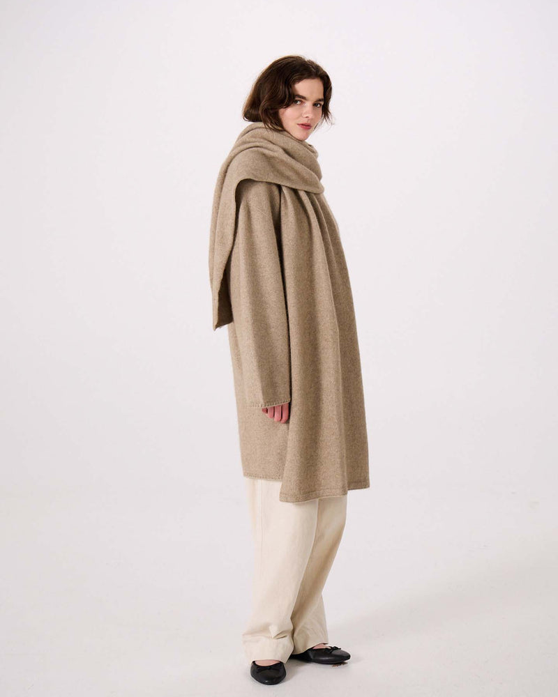 Person wearing a long beige coat with the Umbra Scarf – Natural draped around their neck, paired with cream pants and black shoes, standing against a plain white background. The coat’s elegant silhouette highlights its 97cm long design, offering both style and comfort.