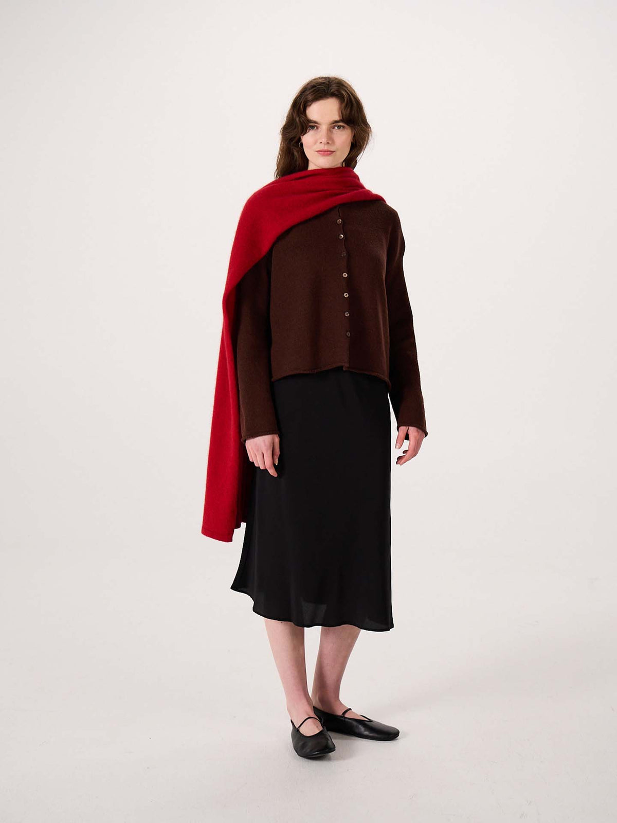 A woman stands against a plain background wearing a brown button-up top, a black skirt, black flats, and the Umbra Scarf – Poppy—a red scarf draped over her shoulder measuring 97cm long and 22cm wide.