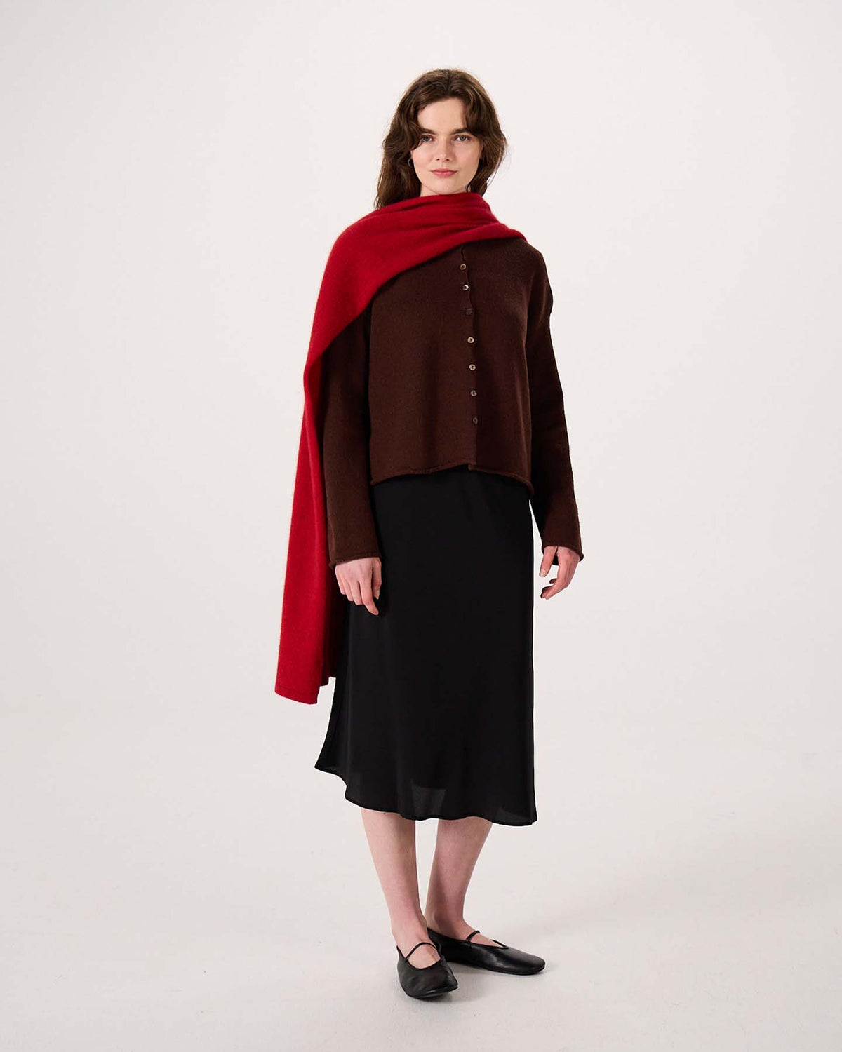 A woman stands against a plain background wearing a brown button-up top, a black skirt, black flats, and the Umbra Scarf – Poppy—a red scarf draped over her shoulder measuring 97cm long and 22cm wide.