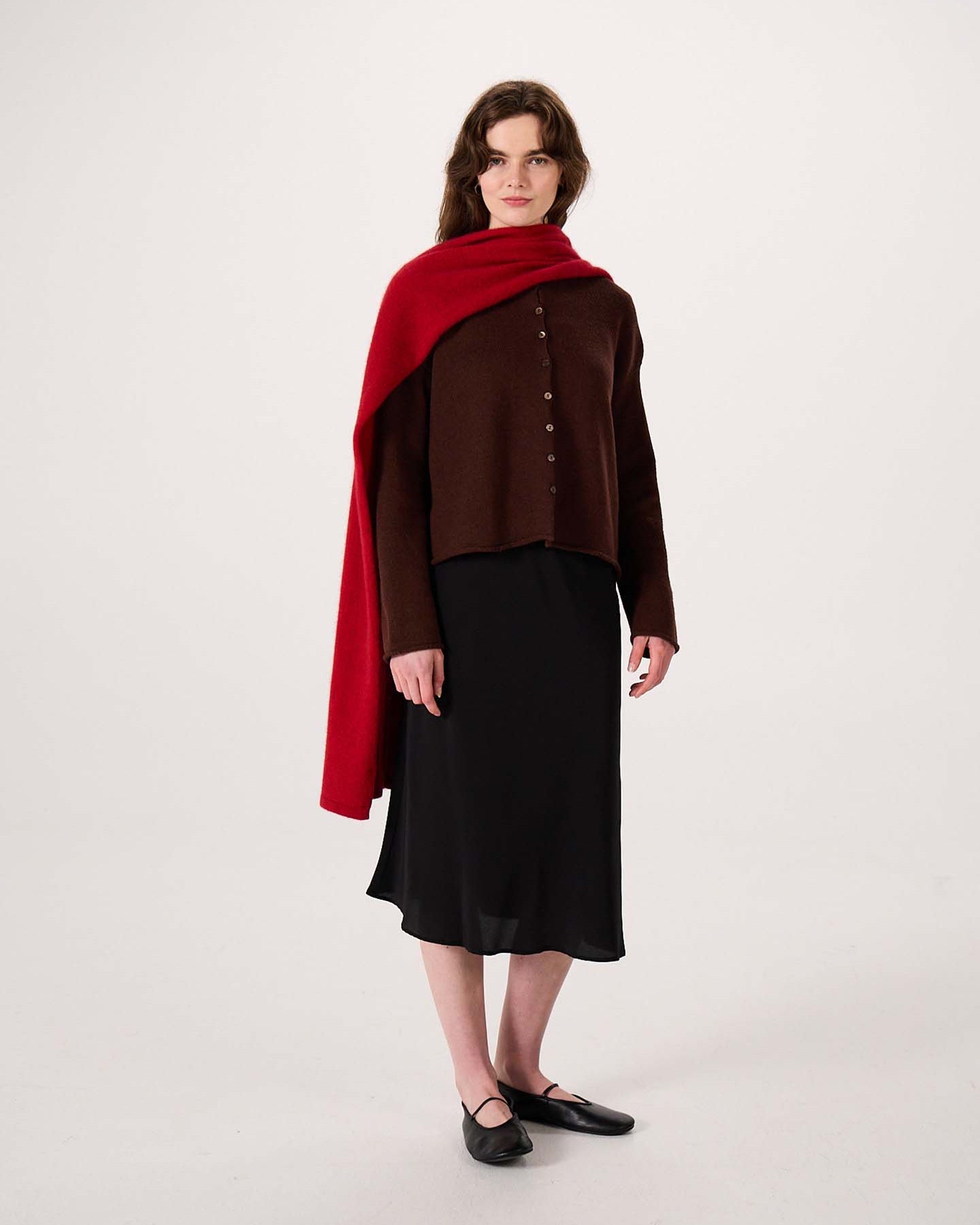 A woman stands against a plain background, wearing a brown buttoned top, black skirt, black shoes, and the Umbra Scarf – Poppy—a long red scarf draped over her shoulders that measures 22 cm wide by 97 cm long.