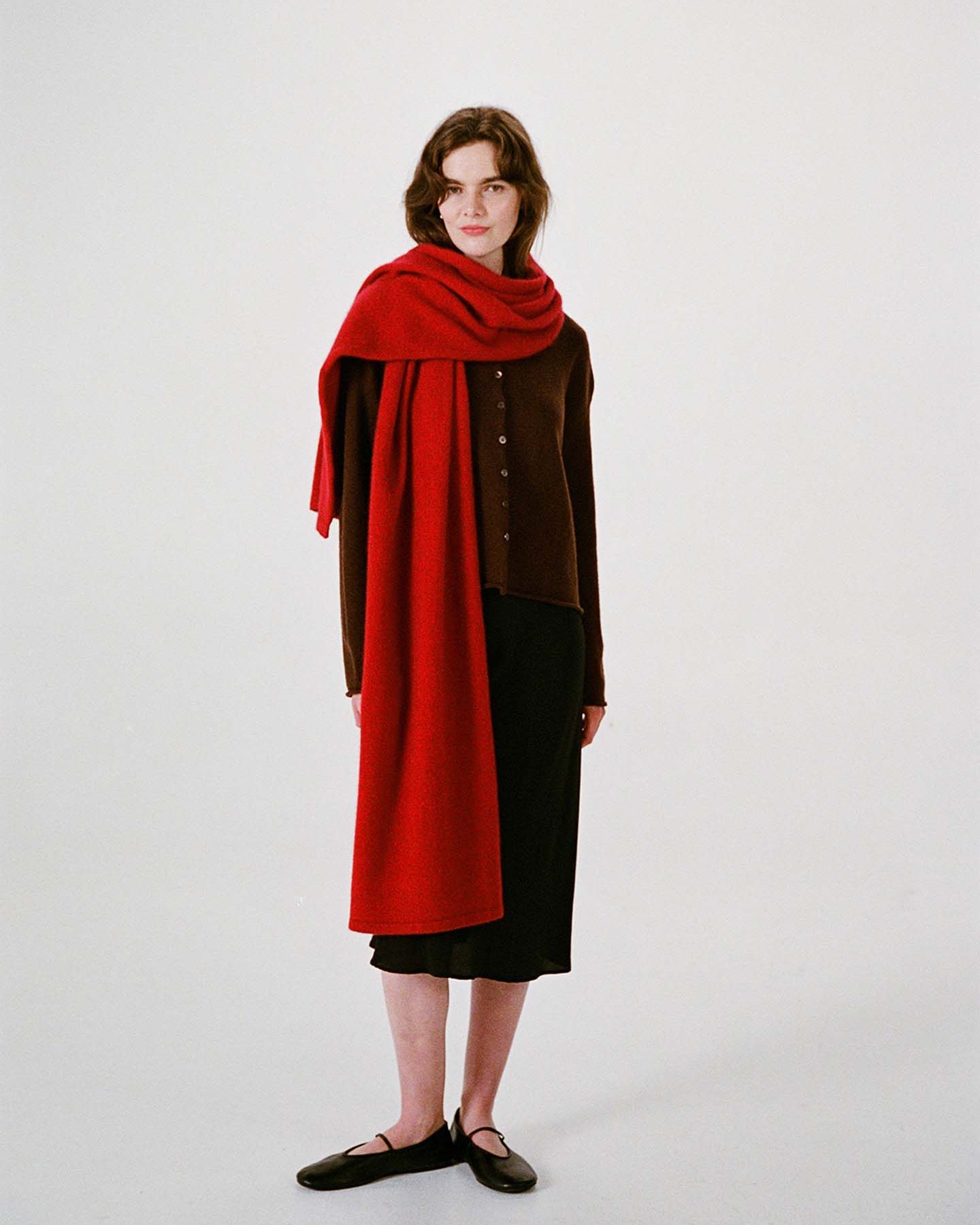 A woman stands against a plain background, wearing a brown buttoned top, black skirt, black shoes, and the Umbra Scarf – Poppy—a long red scarf draped over her shoulders that measures 22 cm wide by 97 cm long.