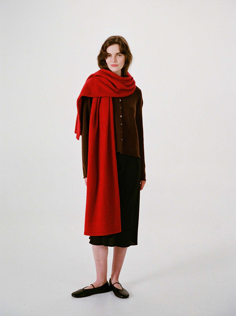 A woman stands against a plain background, wearing a brown buttoned top, black skirt, black shoes, and the Umbra Scarf – Poppy—a long red scarf draped over her shoulders that measures 22 cm wide by 97 cm long.